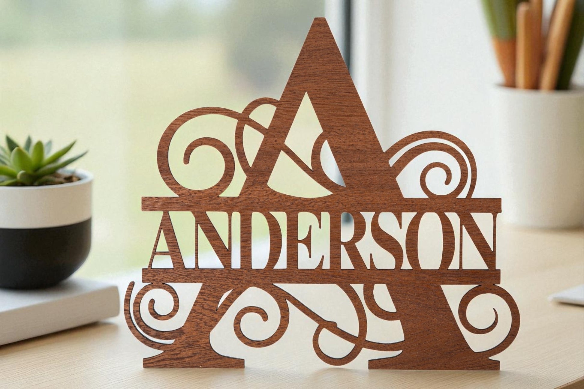 The Custom Walnut Name Cutout by Mirabilia Boutique adds a touch of elegance to any desk, featuring your personalized name with intricate detailing—perfect decor beside a potted plant and window.