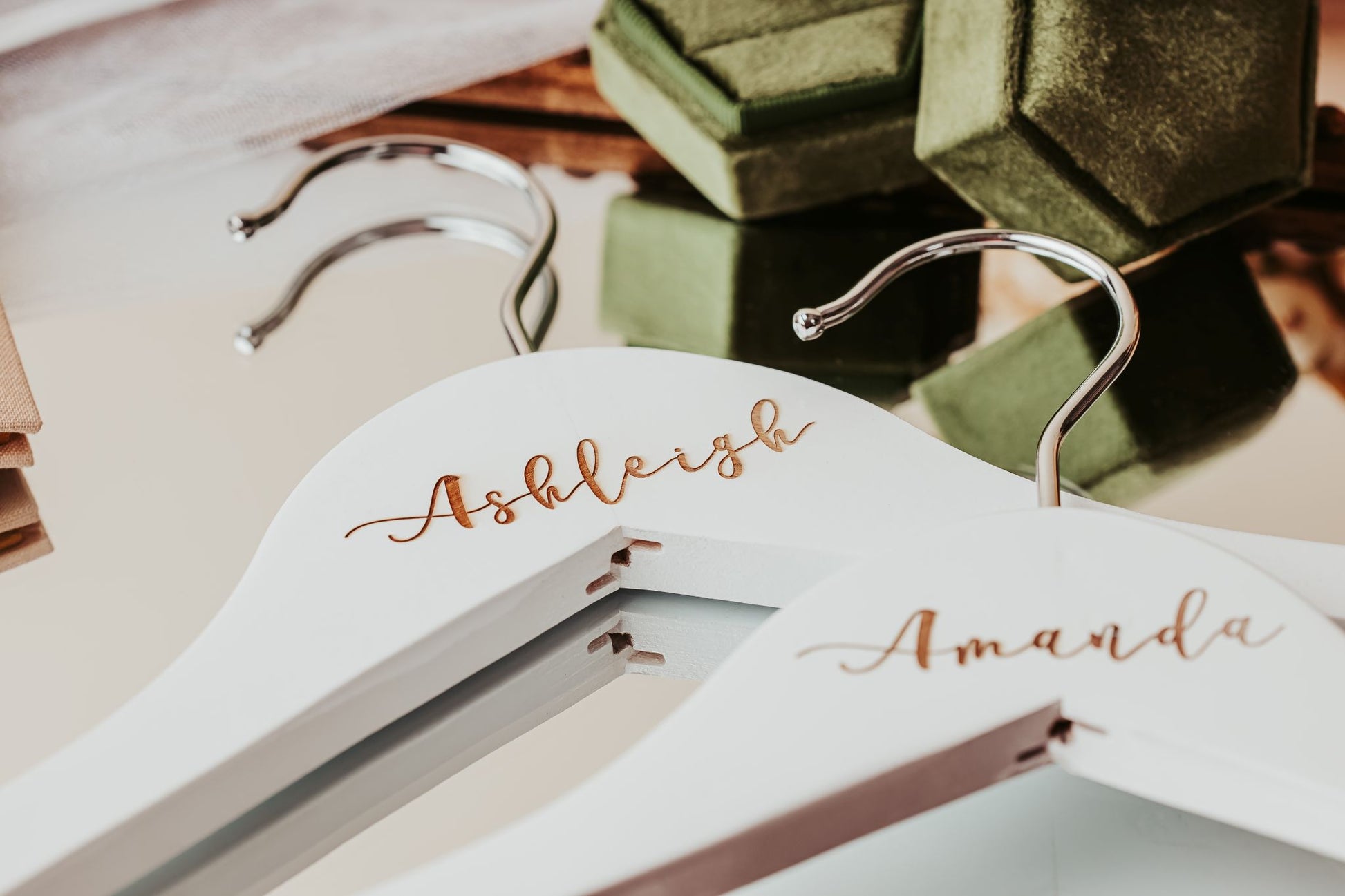 The Personalized Wedding Hangers | Custom Name Hanger Set by Mirabilia Boutique, featuring gold script names, rest on a mirror and add a special touch to wedding day details, with green velvet ring boxes visible in the background.