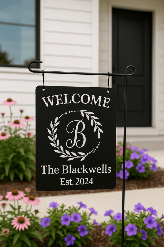 The Mirabilia Boutique Personalized Welcome Garden Sign features white text—"WELCOME, B, The Blackwells, Est. 2024"—and hangs at a white house among blooming purple and pink flowers, making it an ideal custom family yard decor gift.