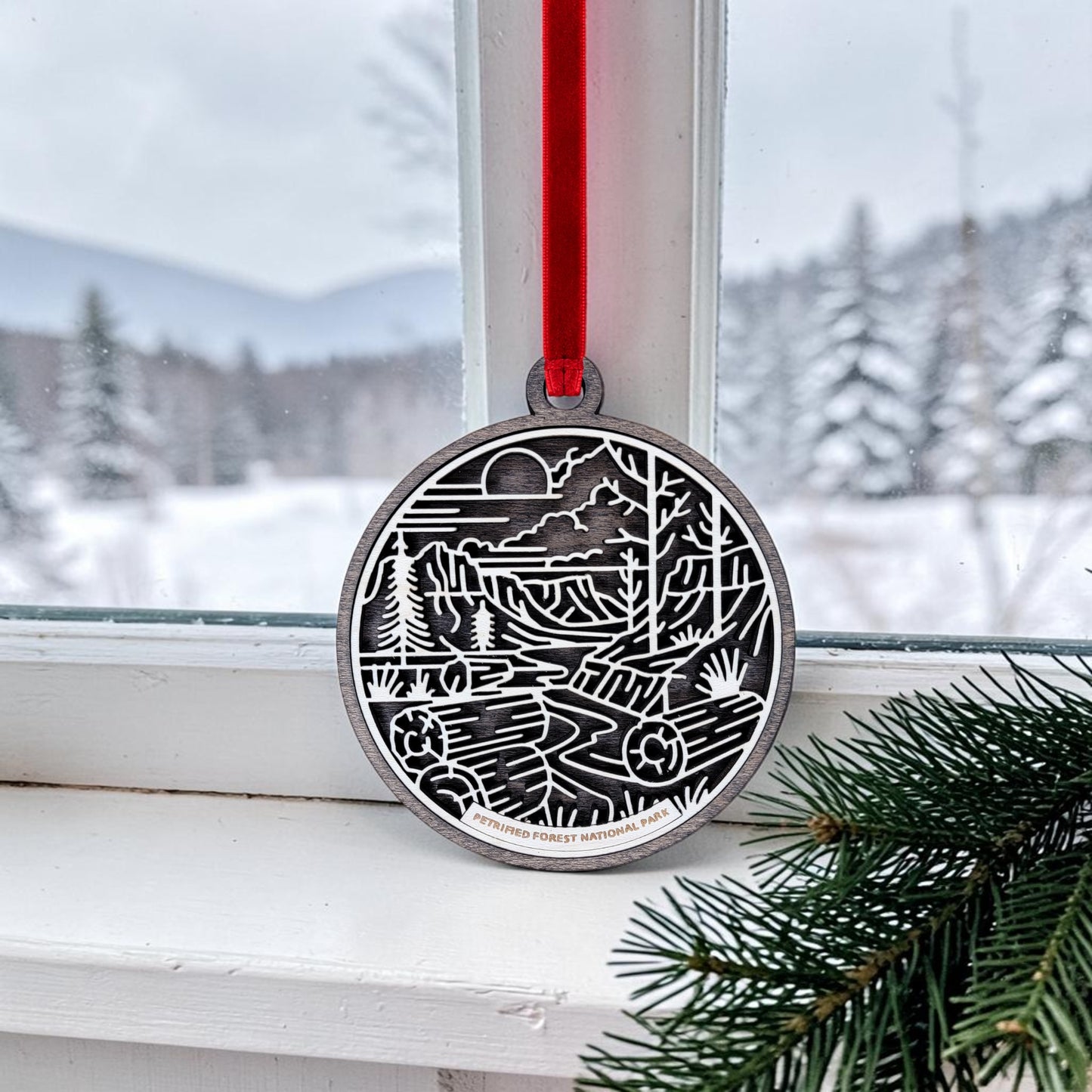 The Petrified Forest National Park Line Art Christmas Ornament by Mirabilia Boutique hangs by a red ribbon on a window, displaying a round wooden design featuring a nature scene; snowy trees and mountains are visible outside, with evergreen branches in the corner.