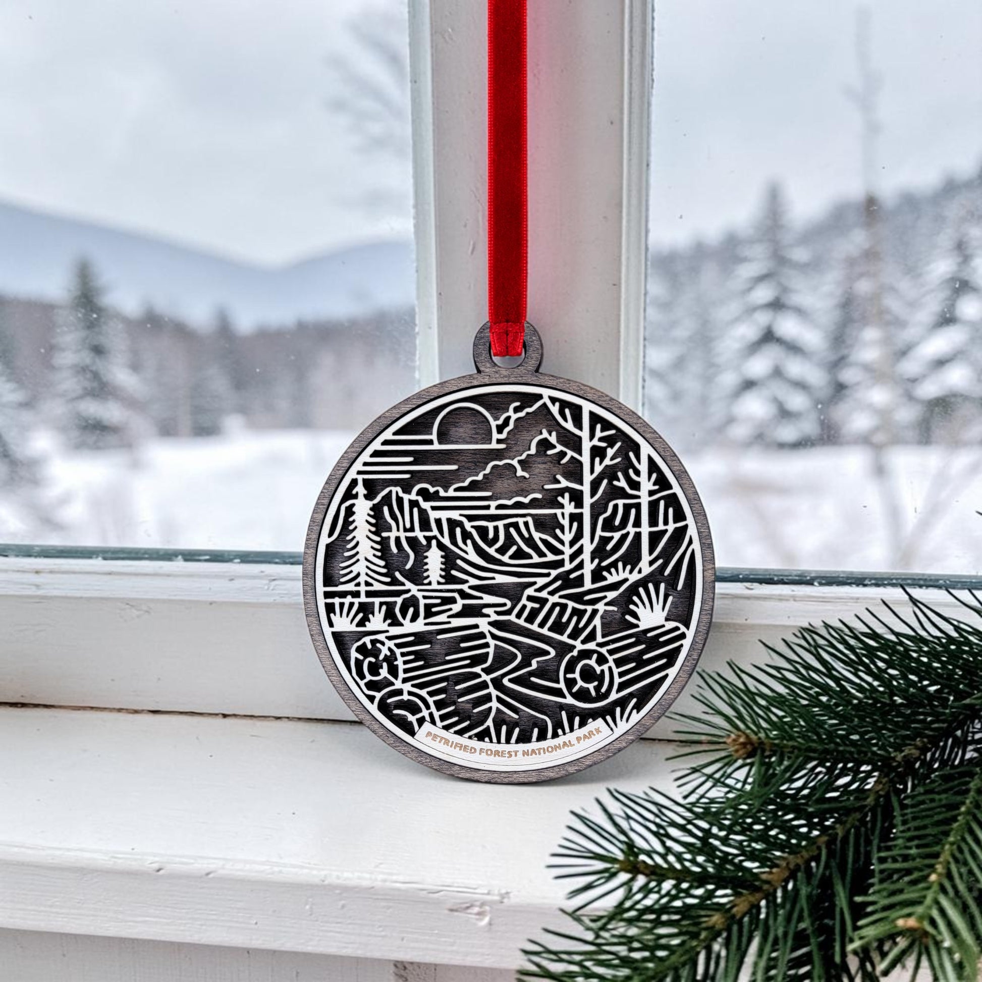 The Petrified Forest National Park Line Art Christmas Ornament by Mirabilia Boutique hangs by a red ribbon on a window, displaying a round wooden design featuring a nature scene; snowy trees and mountains are visible outside, with evergreen branches in the corner.