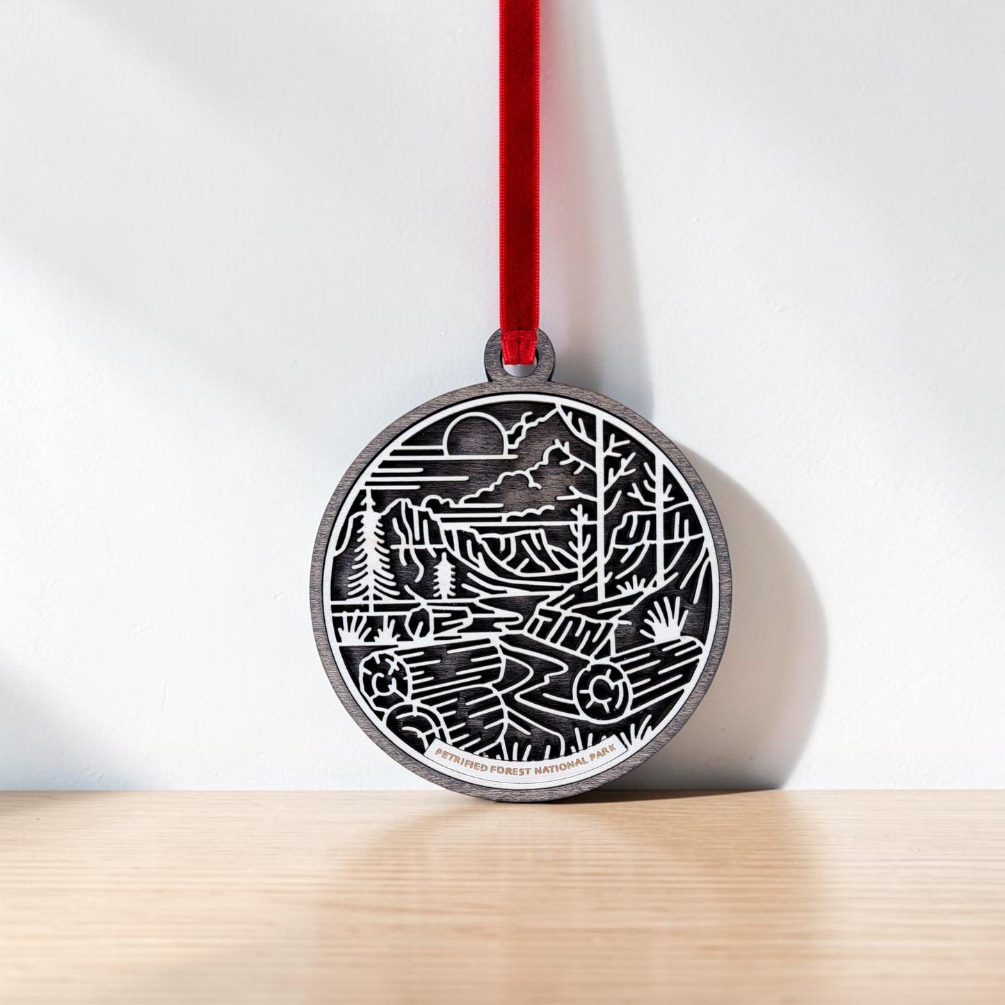 The Petrified Forest National Park Line Art Christmas Ornament by Mirabilia Boutique is an oval-shaped wooden decoration adorned with a red ribbon and features a black and white line art nature scene of mountains, trees, and a river. The ornament rests against a white wall on a light wooden surface.