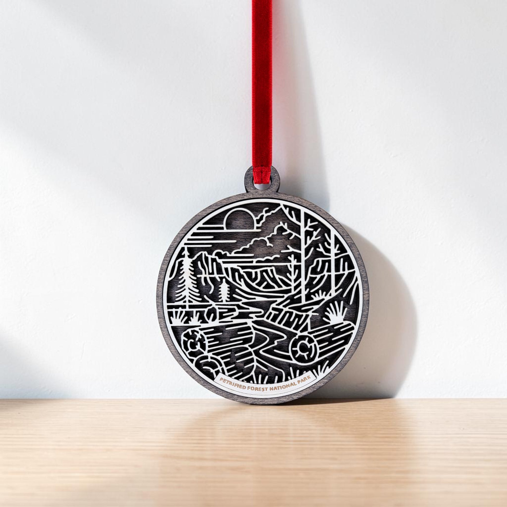 The Petrified Forest National Park Line Art Christmas Ornament by Mirabilia Boutique is an oval-shaped wooden decoration adorned with a red ribbon and features a black and white line art nature scene of mountains, trees, and a river. The ornament rests against a white wall on a light wooden surface.