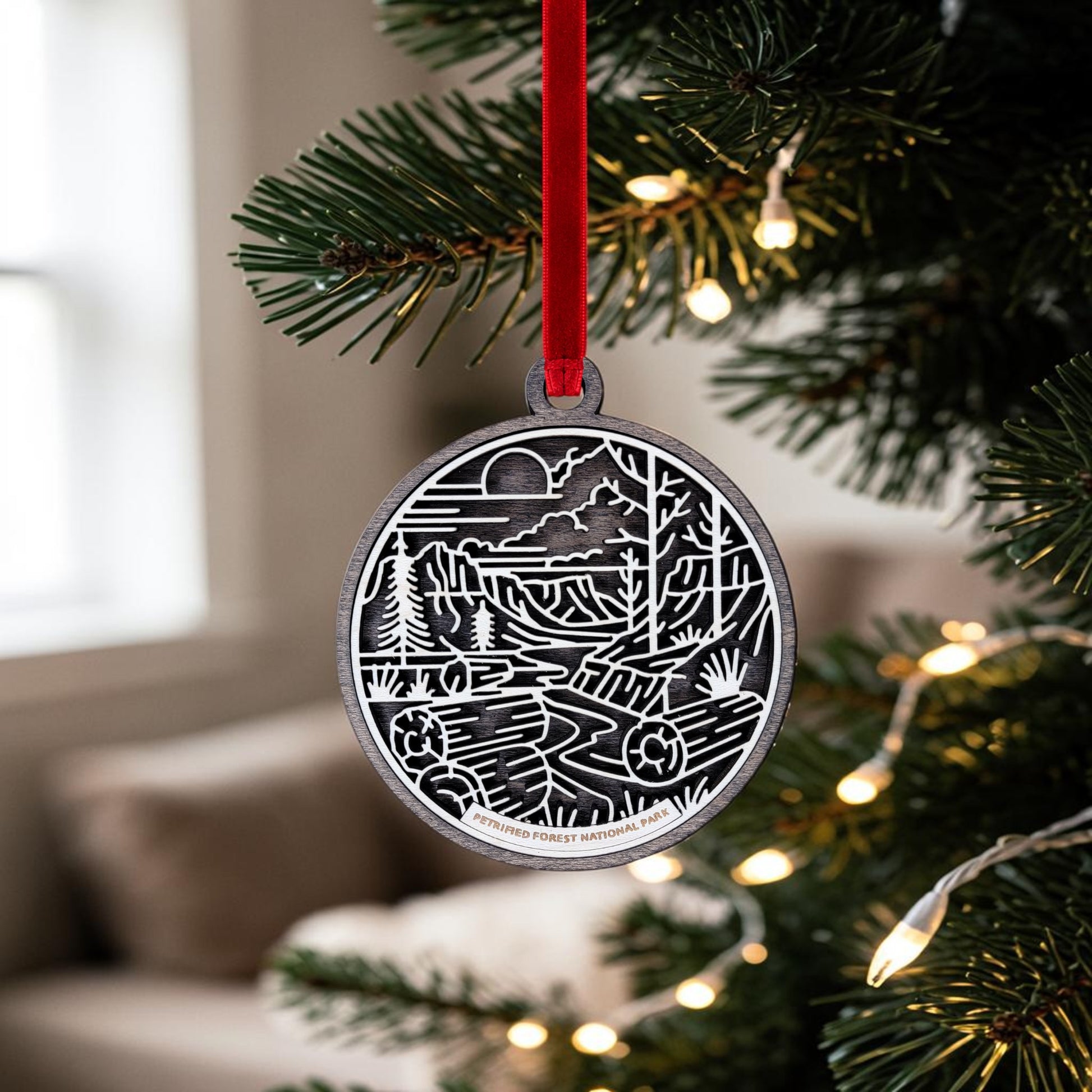 A Petrified Forest National Park Line Art Christmas Ornament by Mirabilia Boutique—a wooden oval ornament showcasing a desert landscape—hangs from a red ribbon on a Christmas tree adorned with white lights. In the background, a softly lit room reveals a window and couch.