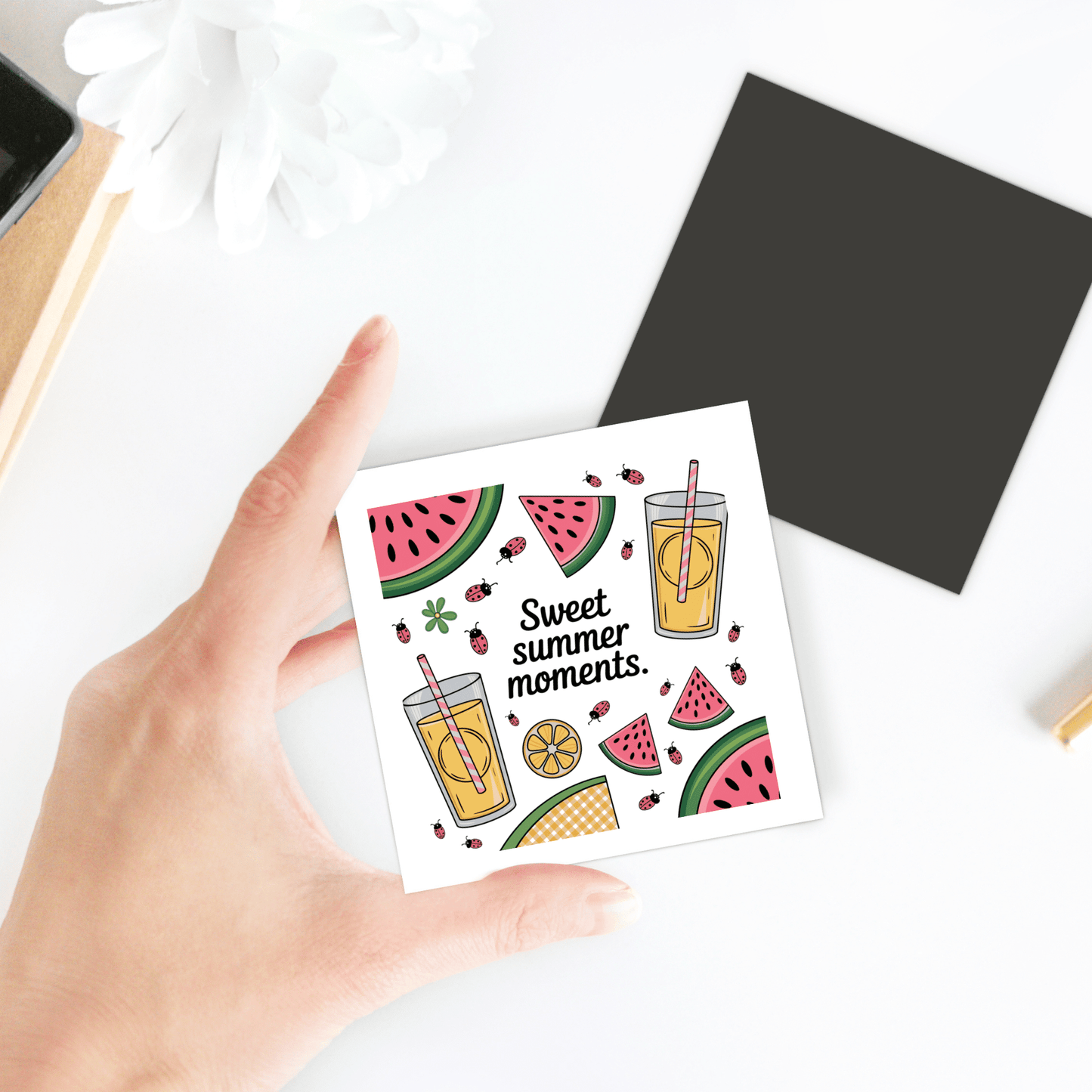 A hand holds the "Sweet Summer Moments Magnet – Watermelon & Lemonade Fridge Art" by Mirabilia Boutique, featuring watermelon, lemonade, and ladybugs. In the background: a notebook, plant, and black square.