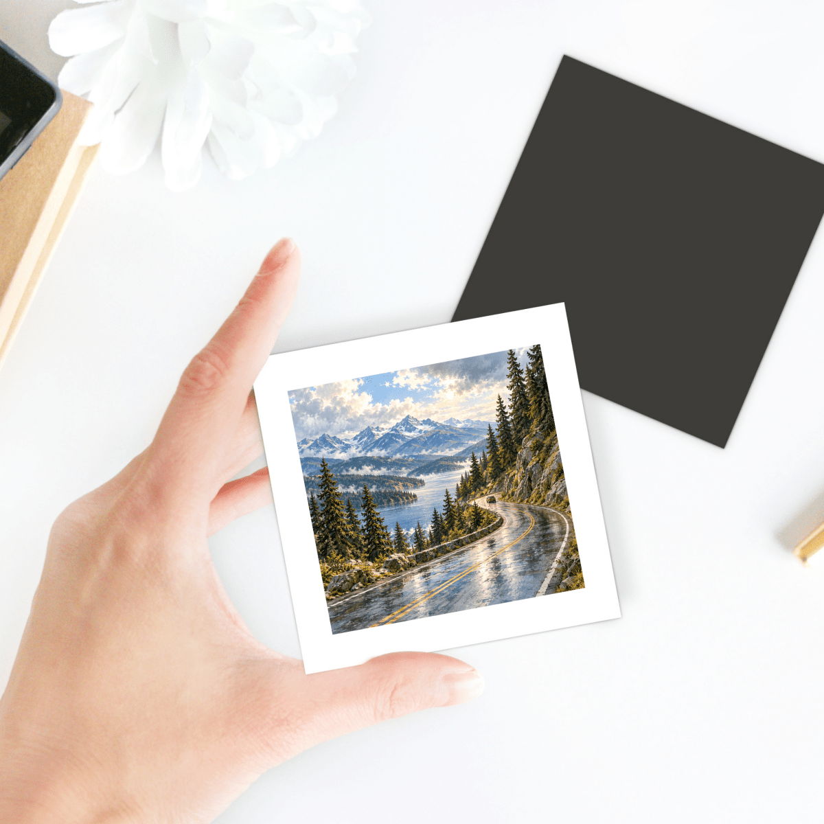 A hand holds The Scenic Byway Magnet, a square mountain landscape featuring a painting of a winding road along a lake and snow-capped peaks.