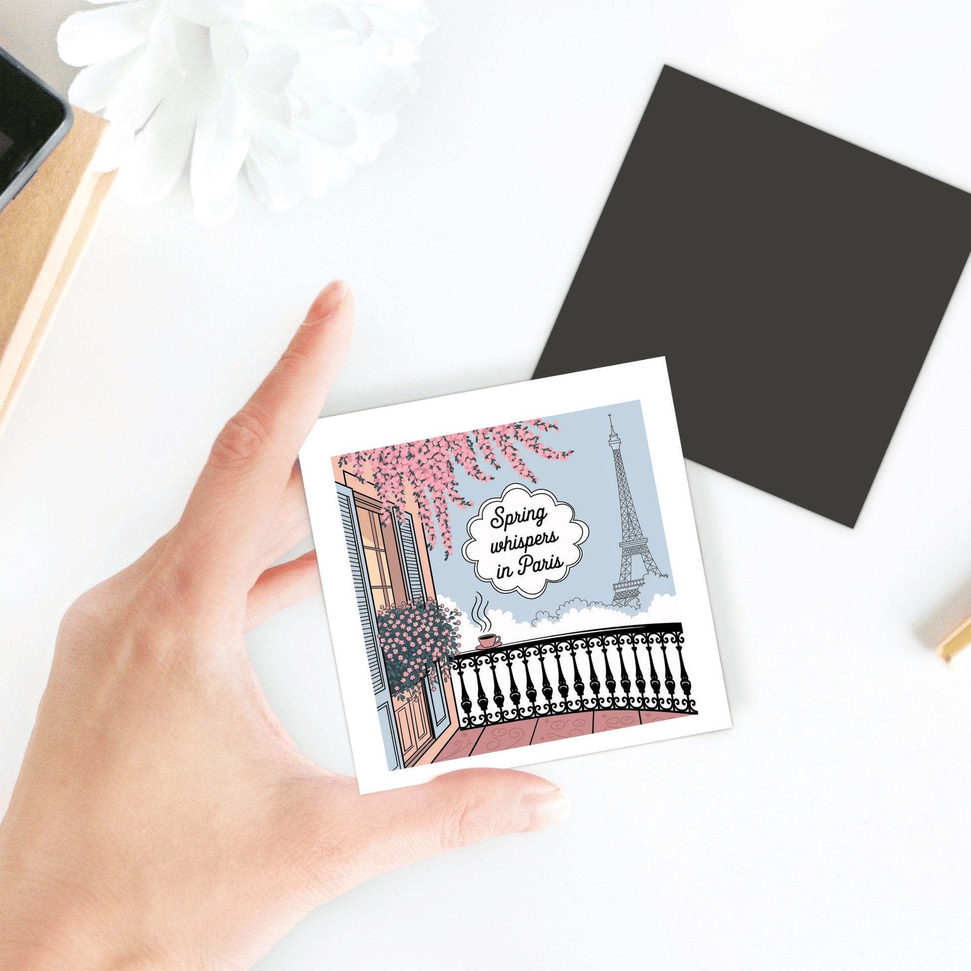 A hand holds the "Spring Whispers in Paris Magnet – Eiffel Tower View" by Mirabilia Boutique, featuring a Paris balcony with flowers, coffee, and the Eiffel Tower—perfect as a spring-inspired gift or Eiffel Tower memento.