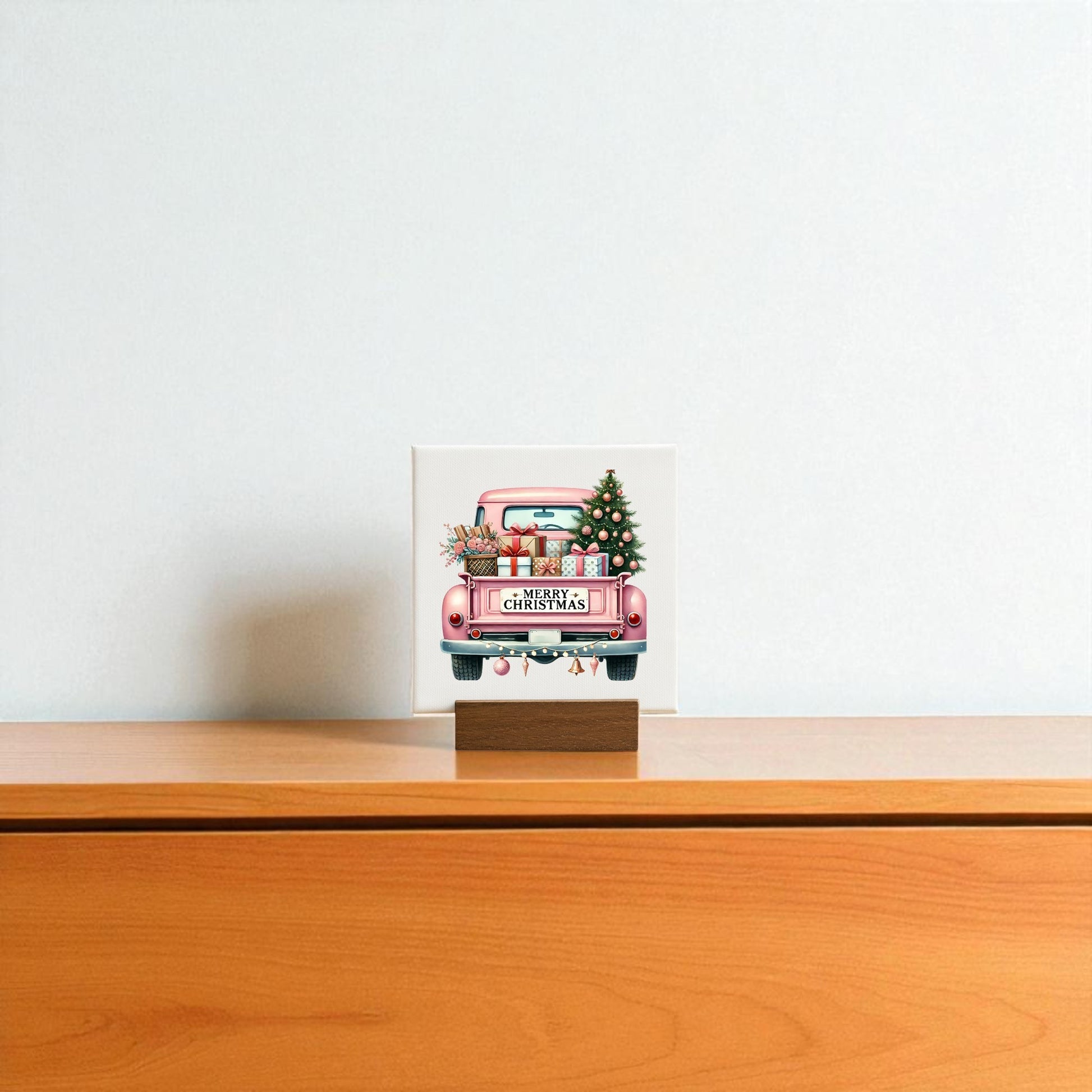 The Pink Christmas Truck Mini Sign by Mirabilia Boutique, featuring gifts and a tree, stands on a wooden base atop a light cabinet, bringing glam holiday decor to the plain white wall.