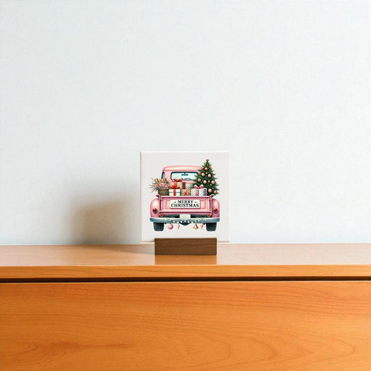 The Pink Christmas Truck Mini Sign by Mirabilia Boutique, featuring gifts and a tree, stands on a wooden base atop a light cabinet, bringing glam holiday decor to the plain white wall.