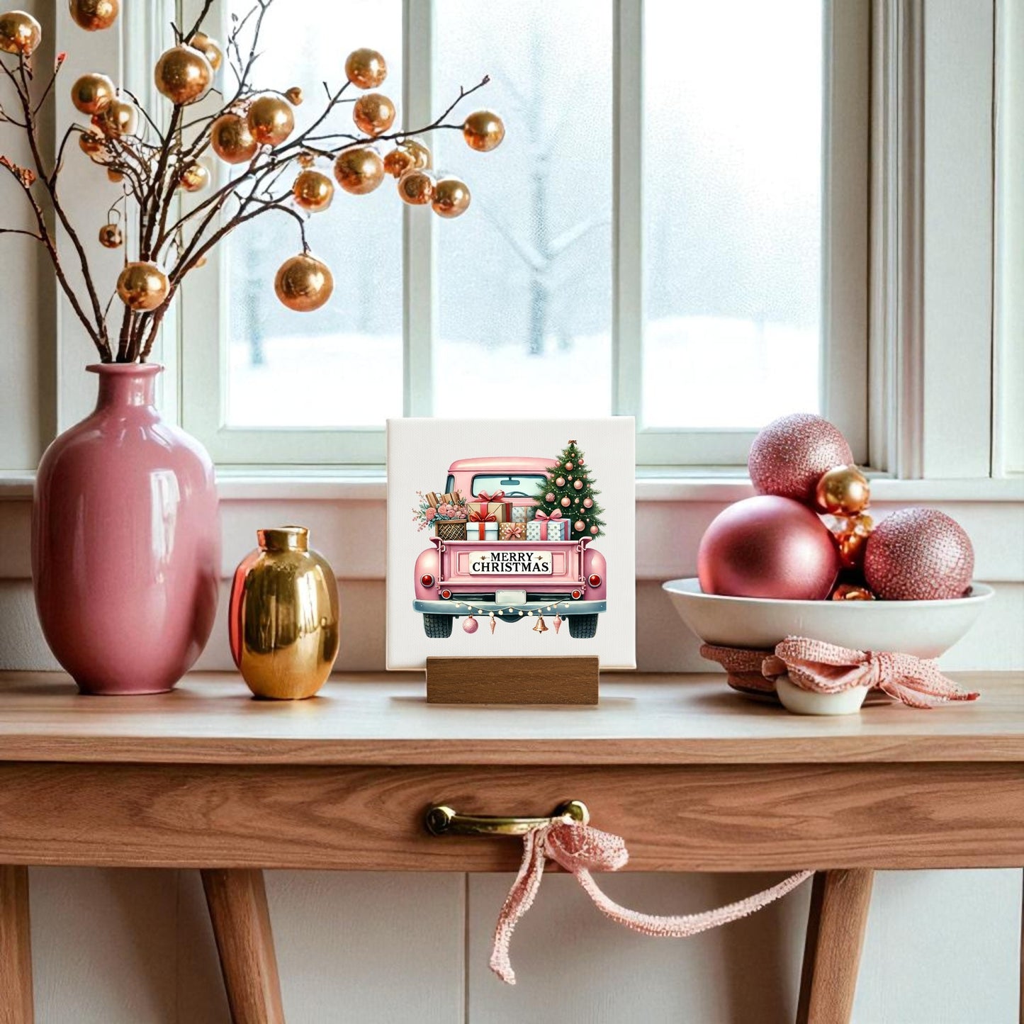 A wooden table decorated with pink and gold ornaments, a pink vase, and a bowl of baubles is topped with the Mirabilia Boutique Pink Christmas Truck Mini Sign. Snow falls outside, completing the glam holiday scene.