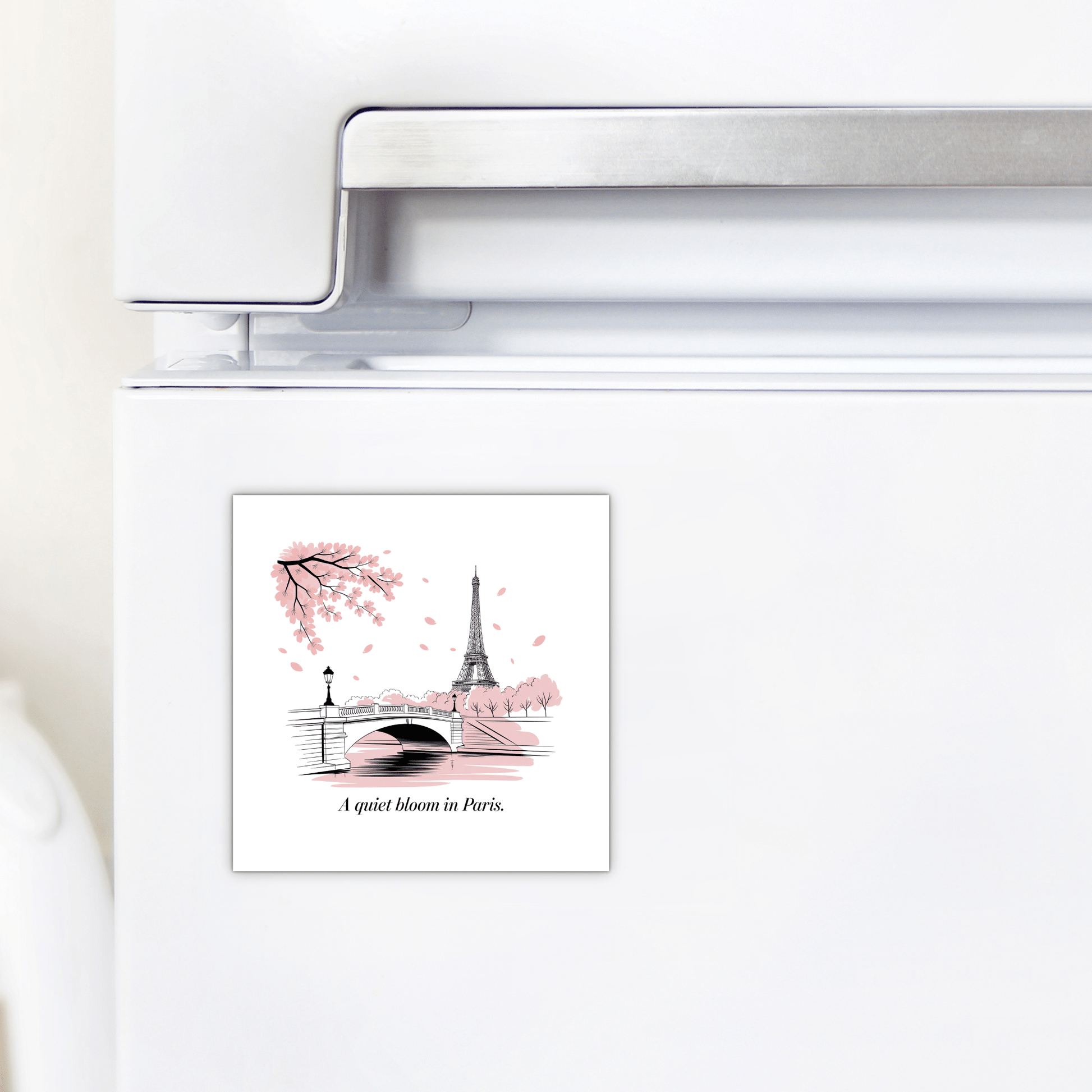 The Mirabilia Boutique “A Quiet Bloom in Paris – Cherry Blossom Magnet” decorates a fridge with a square design showing the Eiffel Tower, a bridge, pink-blossomed trees, and the phrase “A quiet bloom in Paris.”.