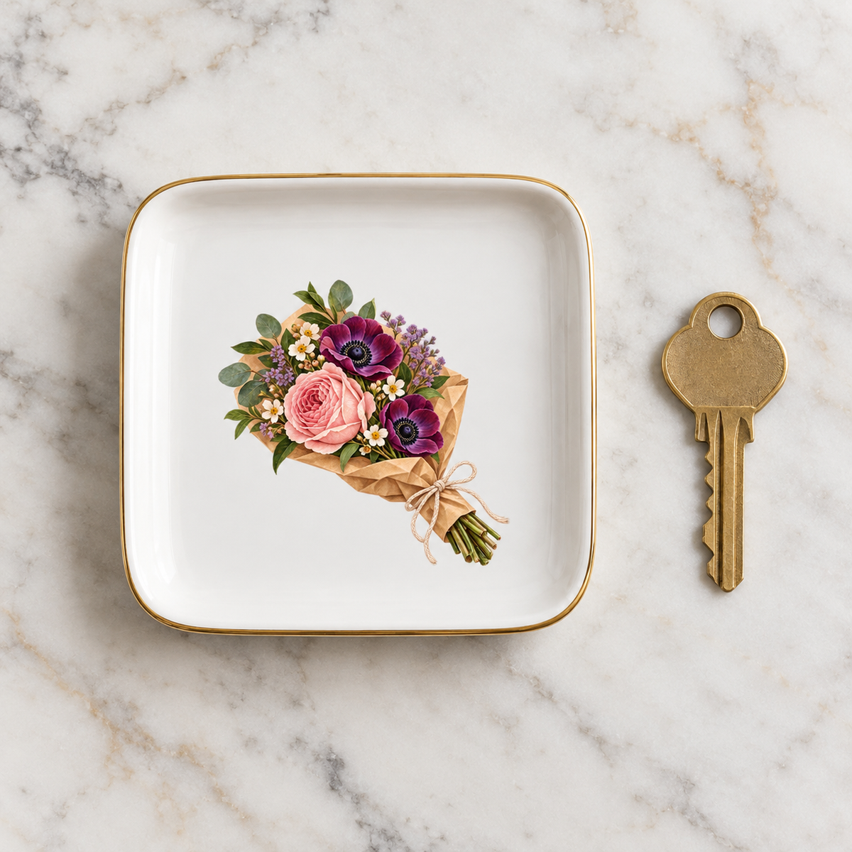 The Florist’s Parcel Jewelry Tray by Mirabilia Boutique is a square white floral dish with a gold rim and a bouquet of pink and purple flowers, sitting on a marble surface next to a brass key.