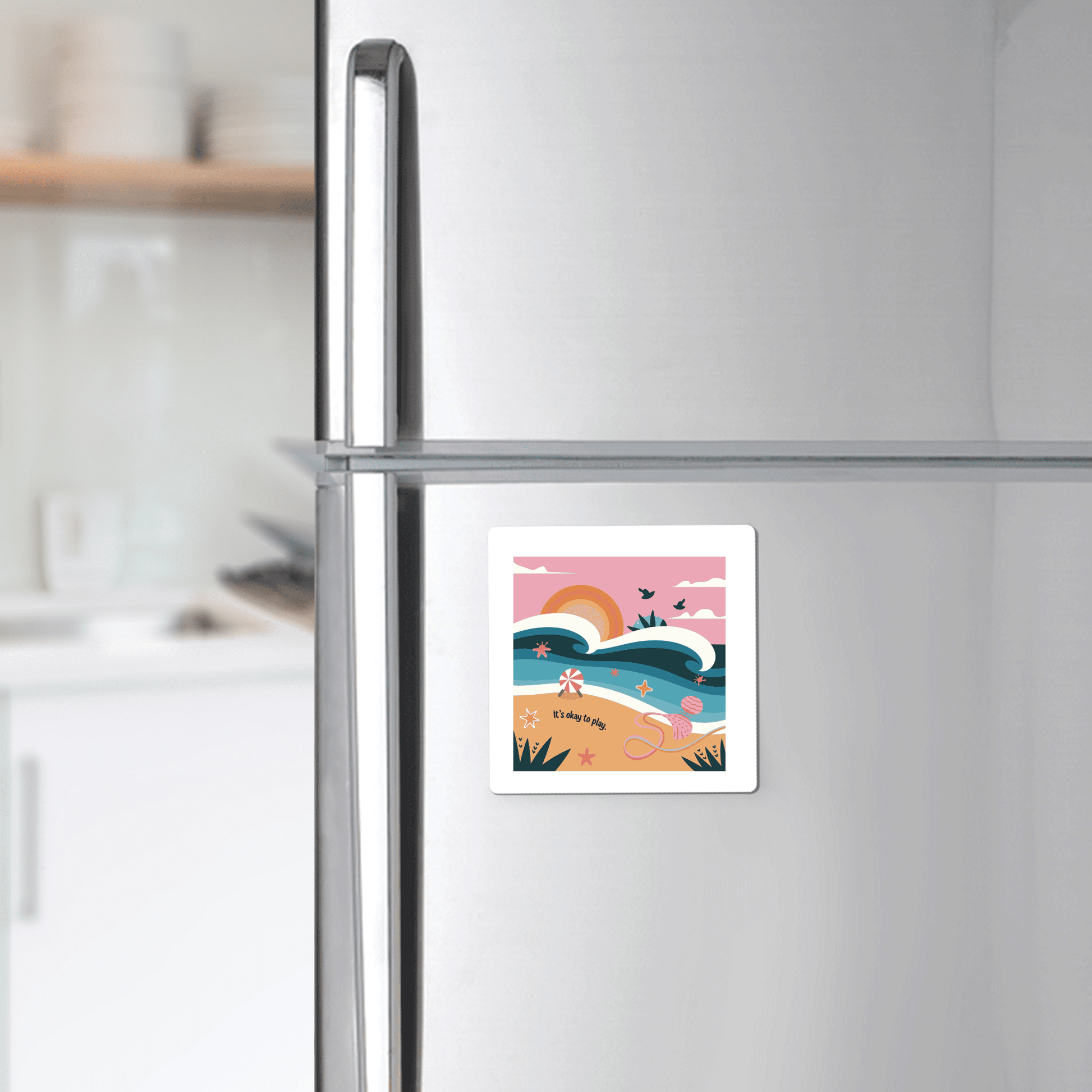 A silver refrigerator in the kitchen displays Mirabilia Boutique’s “It’s Okay to Play Magnet – Whimsical Ocean Joy,” featuring waves, a rainbow, birds, and a pink sky. Shelves with dishes appear in the background.