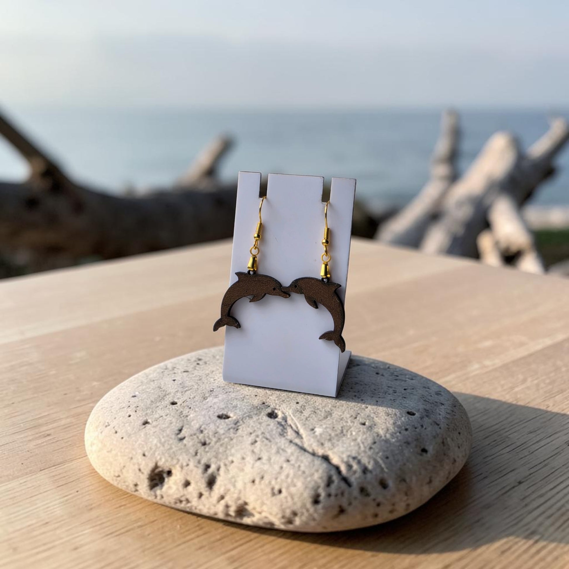 Mirabilia Boutique’s Dolphin Earrings | Lasercut Walnut Wood rest on a white card atop a smooth stone and wood surface, with ocean and driftwood in the background—a perfect ocean-inspired accessory for sea lovers.