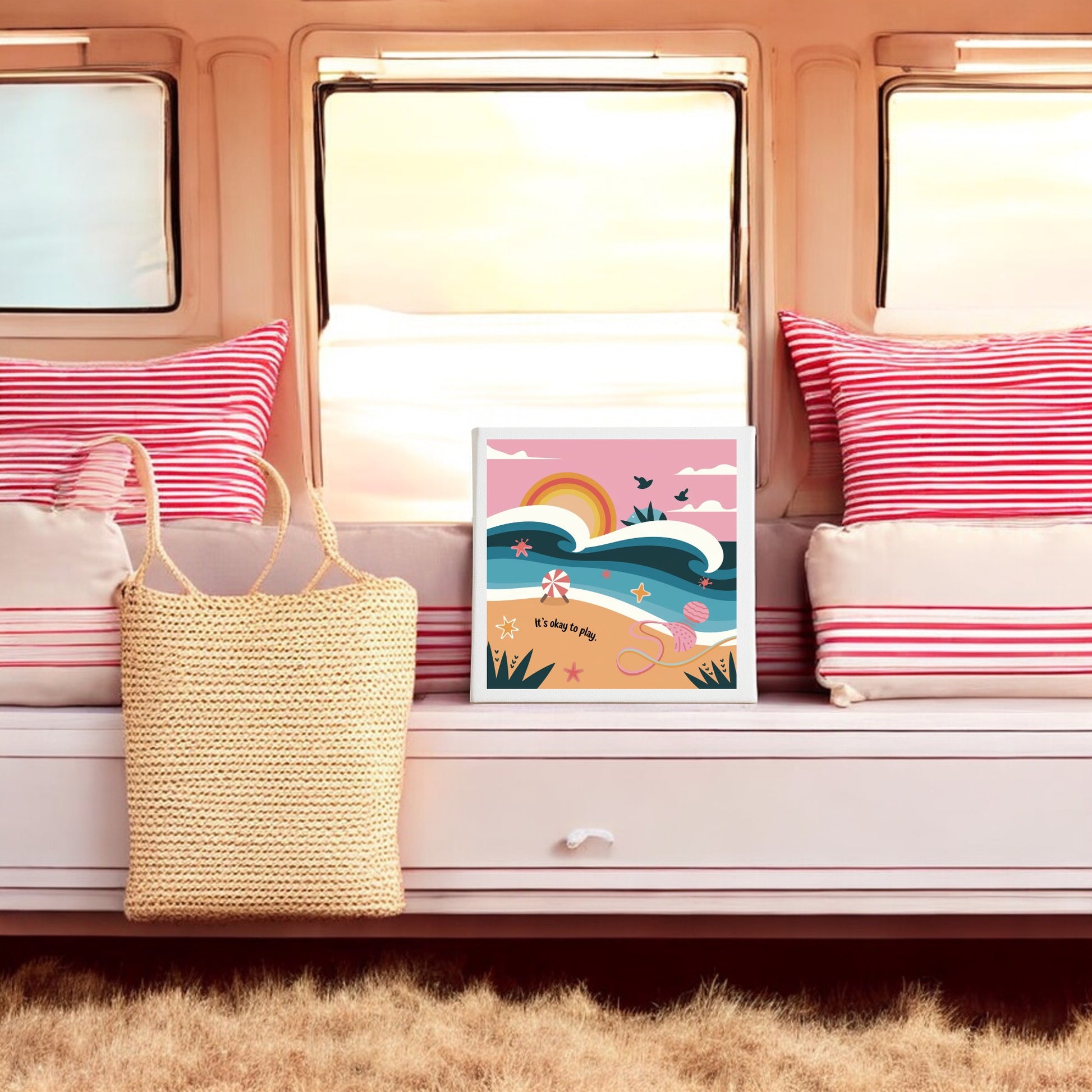A cozy window seat with red and white striped cushions, a woven tote, and the "It’s Okay to Play Canvas – Whimsical Beach Wall Art" by Mirabilia Boutique adds vibrant color and charm against large windows.