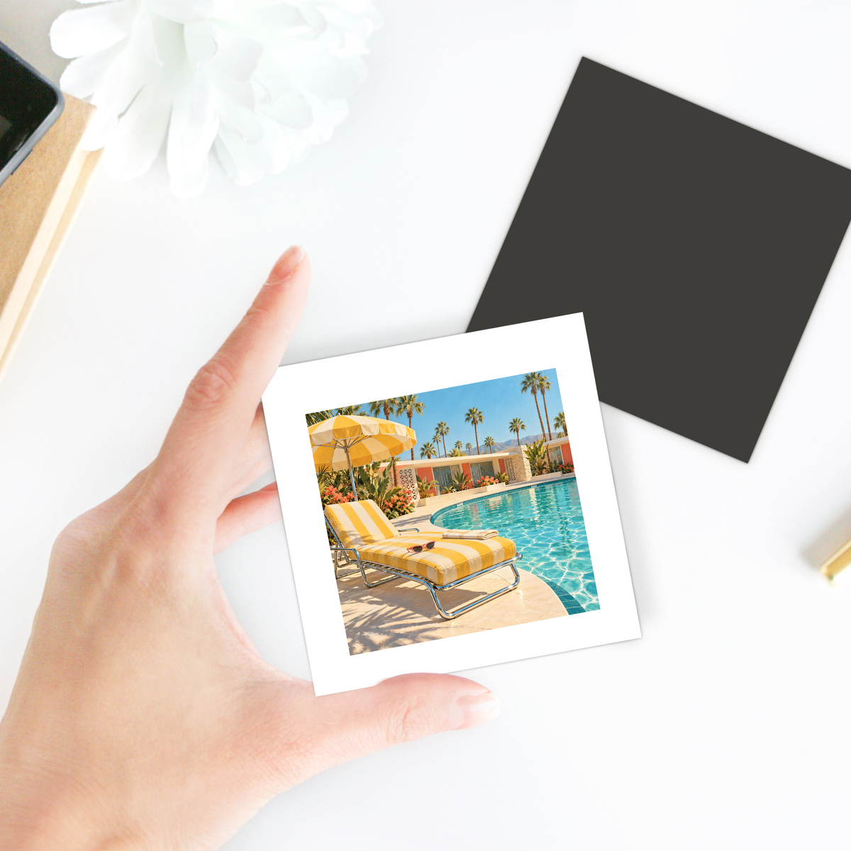 A hand holds a vibrant photo—yellow-striped lounge chairs, palm trees, and blue water—featured on the Afternoon at the Pool Magnet | Chrome & Starlight by Mirabilia Boutique for your fridge collection.