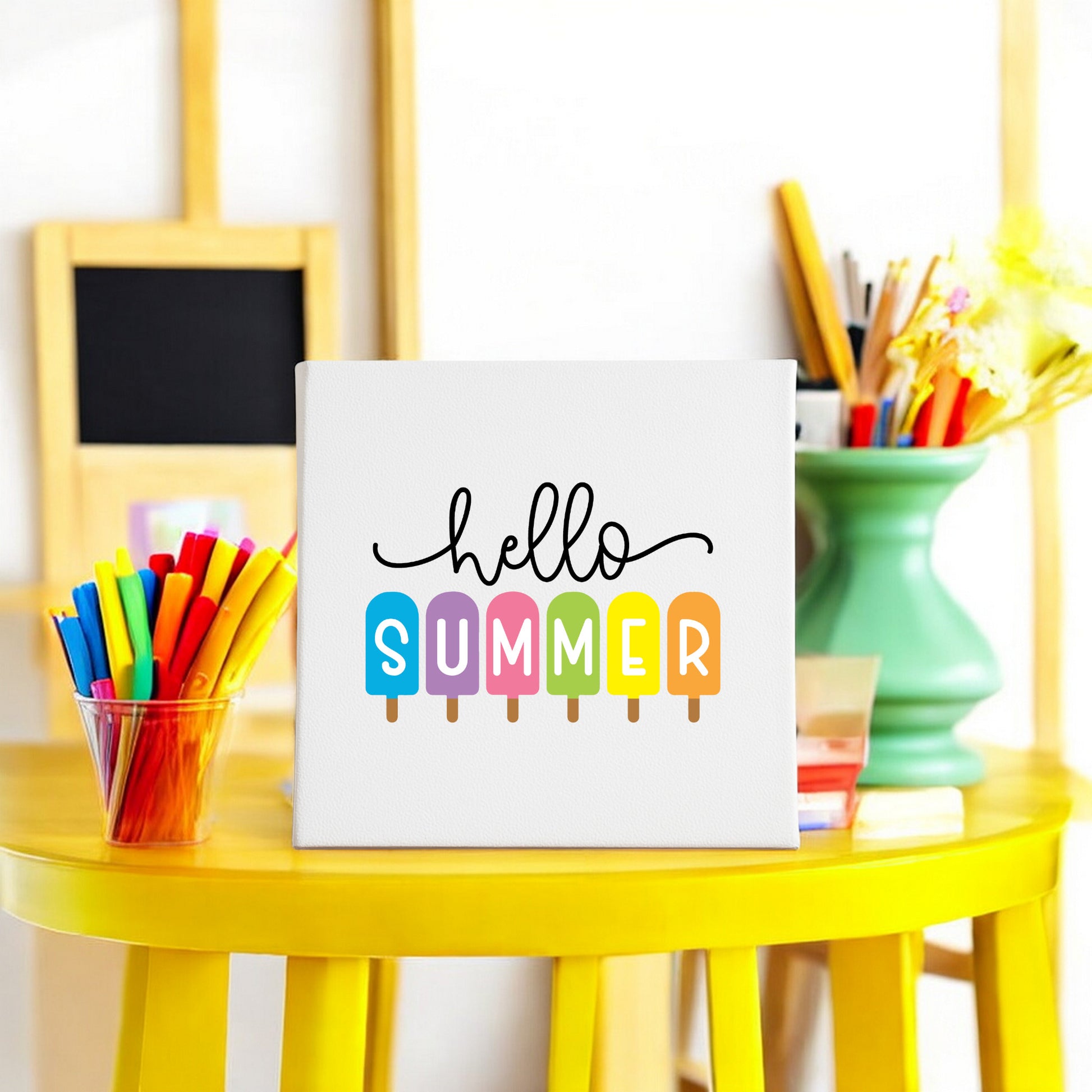 The Mirabilia Boutique Hello Summer Popsicle Canvas Sign (8x8) features “hello summer” with colorful popsicles, making cheerful wall art. Art supplies, a green vase, and a small blackboard in the background enhance the creative vibe.