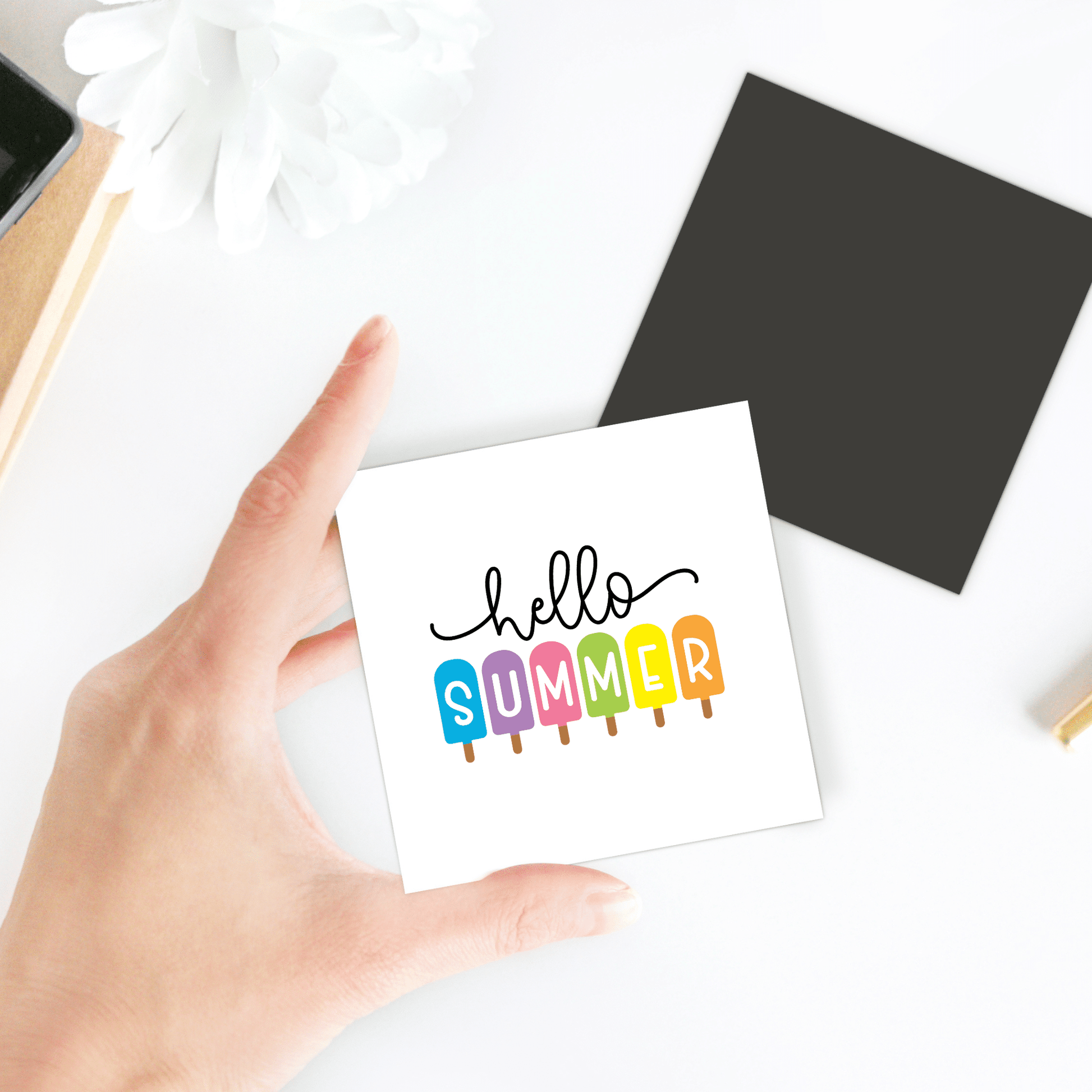 A hand holds the Hello Summer Popsicle Magnet by Mirabilia Boutique, featuring "SUMMER" spelled with rainbow popsicles—a vibrant ice pop fridge art. Nearby: a black square, white flower, and brown object on a white surface.