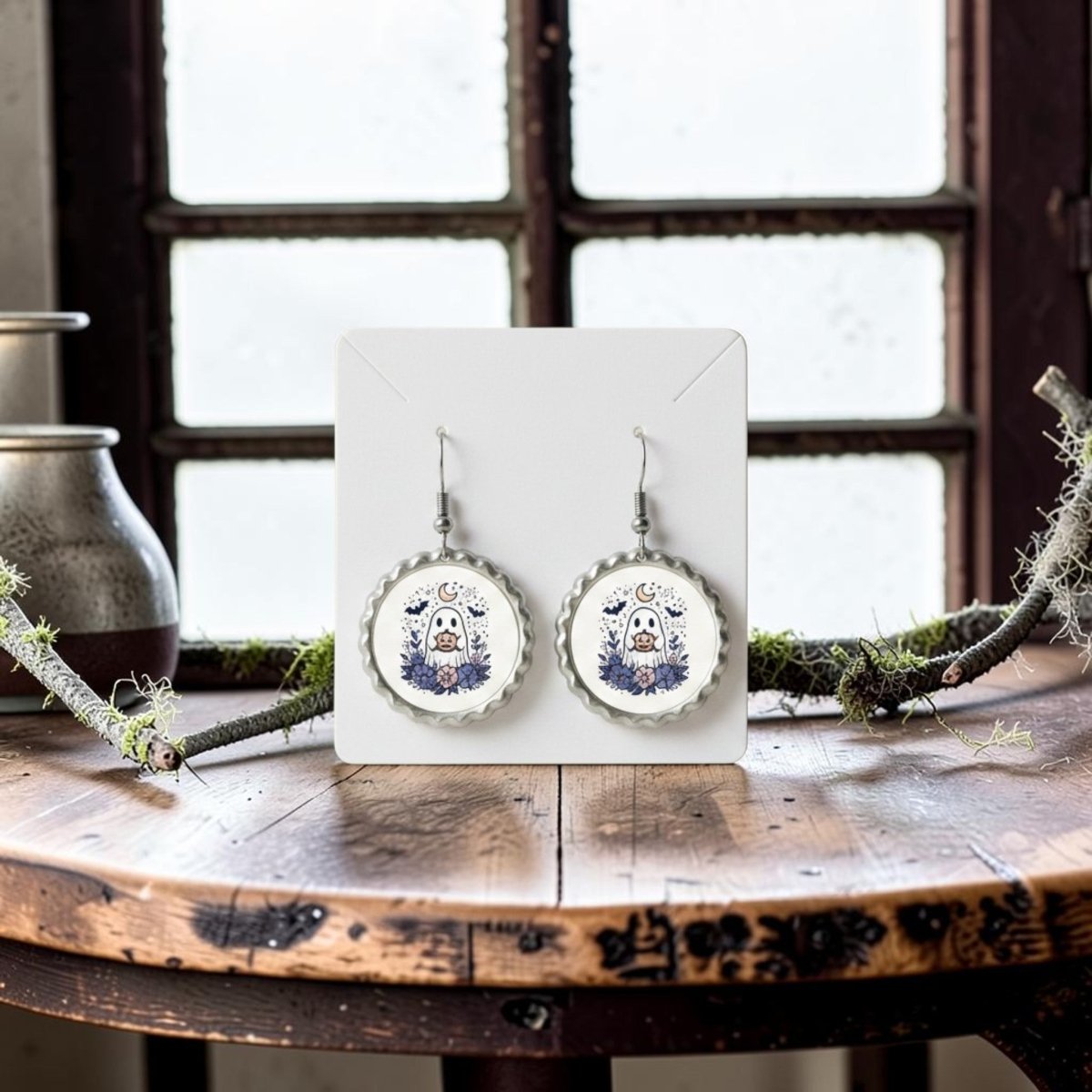 A pair of Crescent Moon Pumpkin Ghost Scalloped Earrings by Mirabilia Boutique, displayed on a card, sits atop a rustic wooden table exuding gothic cottagecore charm, with a metal jug and mossy branches near a window in the background.