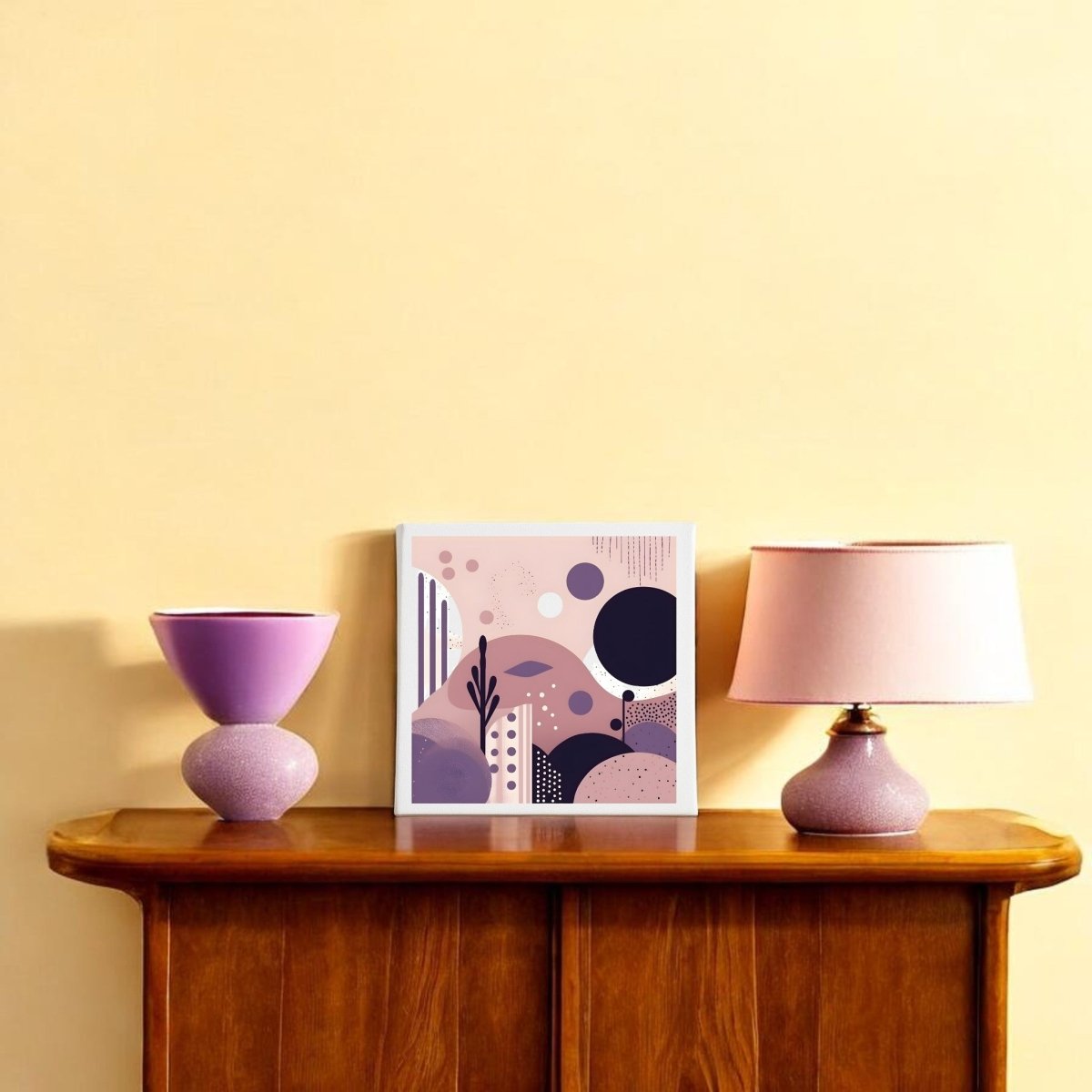 A wooden table against a pale yellow wall holds a pink lampshade lamp, a pink vase, and the Purple Dusk Mid Century Modern Canvas by Mirabilia Boutique—an abstract decor piece in pastel tones with geometric and organic shapes.