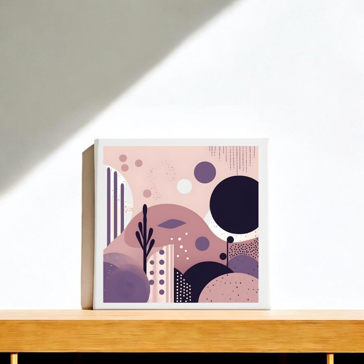 The Purple Dusk Mid Century Modern Canvas by Mirabilia Boutique, featuring abstract geometric shapes in pink, purple, and black, sits on a light wooden shelf with soft sunlight and shadows across the wall and surface.