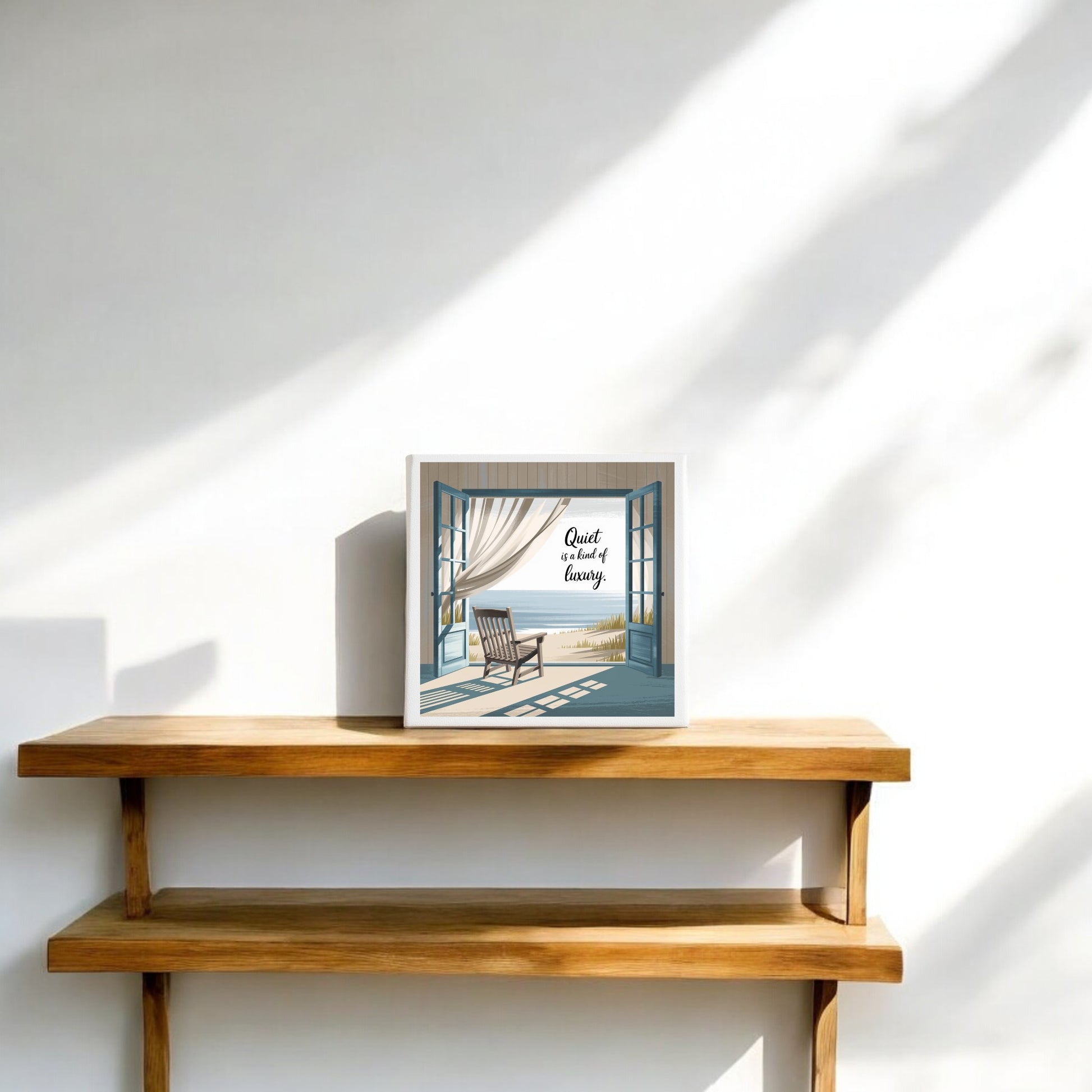 A light wooden shelf displays the "Quiet Is a Kind of Luxury Canvas – Peaceful Summer Wall Art" by Mirabilia Boutique, featuring sunlit blue windows and a rocking chair that echo the quiet luxury style of the Mirabilia Summer collection.