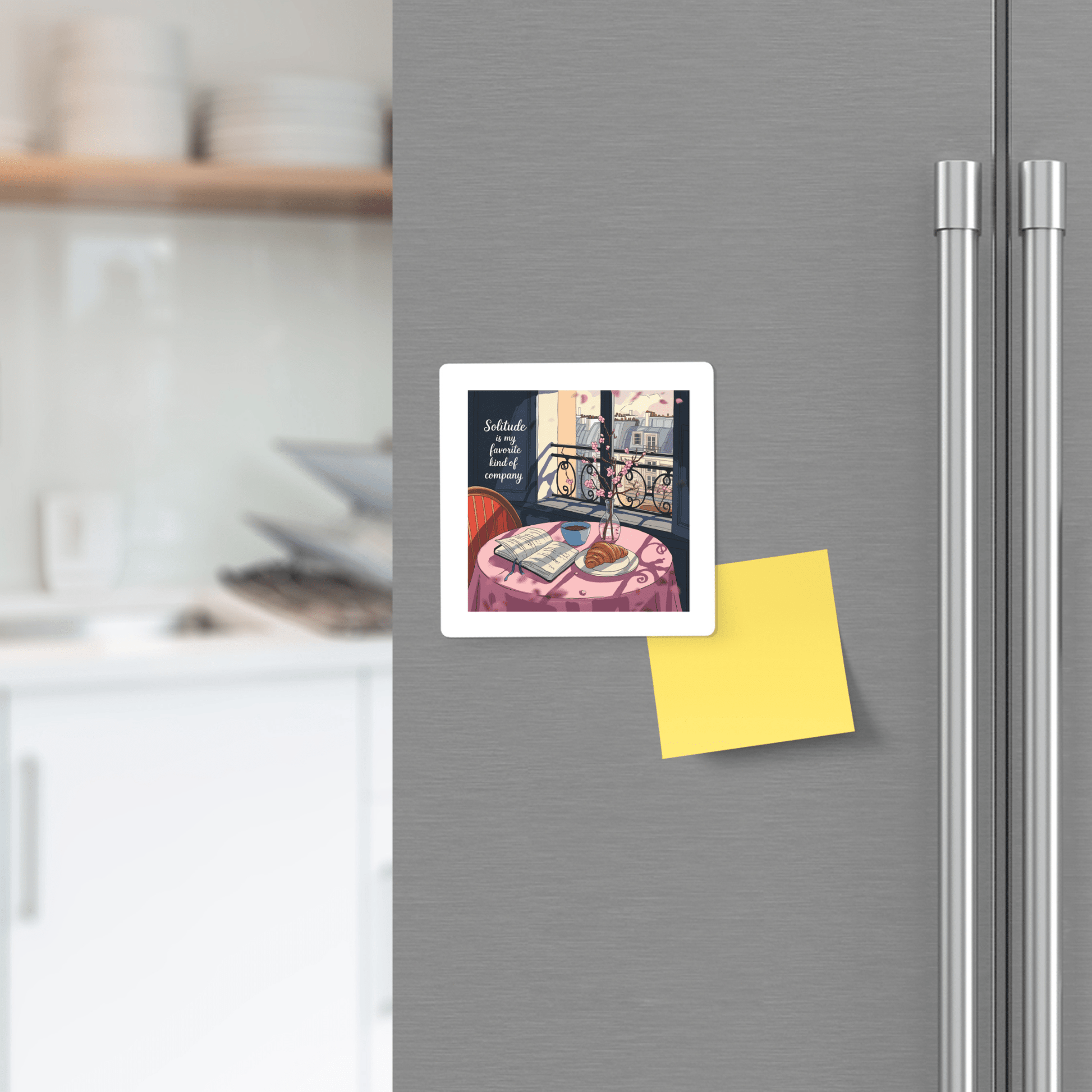 A refrigerator door displays the Solitude Magnet – Paris Morning Still Life by Mirabilia Boutique, featuring a Parisian table scene, above a blank yellow sticky note, with a blurred modern kitchen in the background.