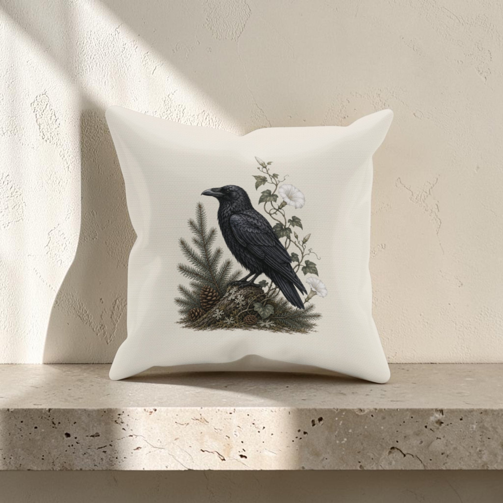 The Quiet Raven Decorative Pillow | 16x16 Woodland Decor from Mirabilia Boutique, showcasing a black raven perched on a pine branch amid delicate white flowers, rests elegantly on a beige stone shelf against a softly textured light wall.