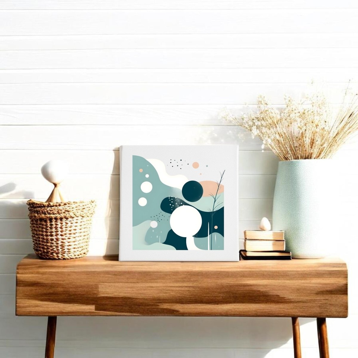 A wooden table against a white wall features Mirabilia Boutique's Quiet Tides Mid Century Modern Canvas, a vase with dried flowers, books, and a woven basket. Sunlight enhances the minimalist, cozy vibe.