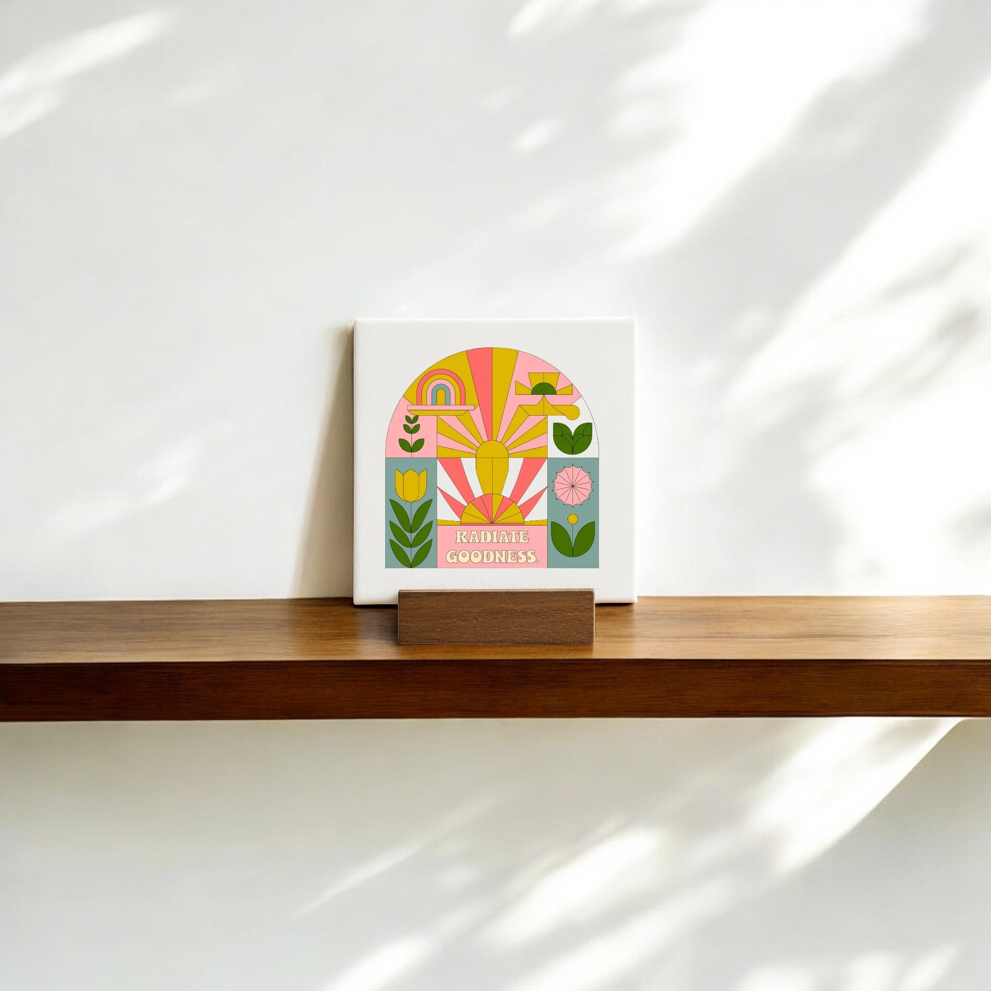 The Mirabilia Boutique "Radiate Goodness" Mini Canvas Sign with Base features colorful abstract flowers and rainbows, adding an encouraging touch to your shelf or desk in soft sunlight against a white wall.