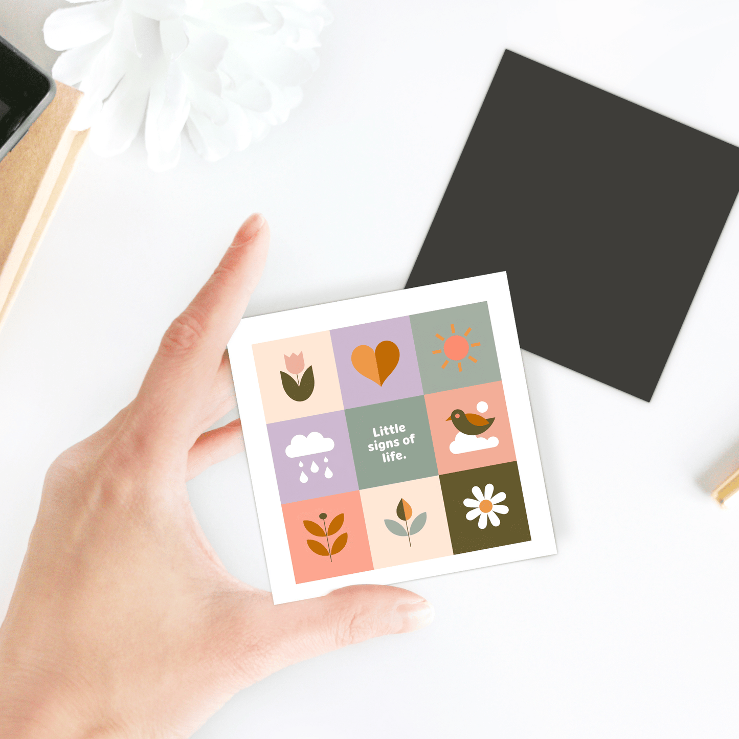 A hand holds the Mirabilia Boutique Little Signs of Life Pastel Grid Magnet, featuring colorful illustrations like a flower, heart, and sun with "Little signs of life" at center. A perfect inspirational gift or minimalist decor accent. Nearby are black and white squares.