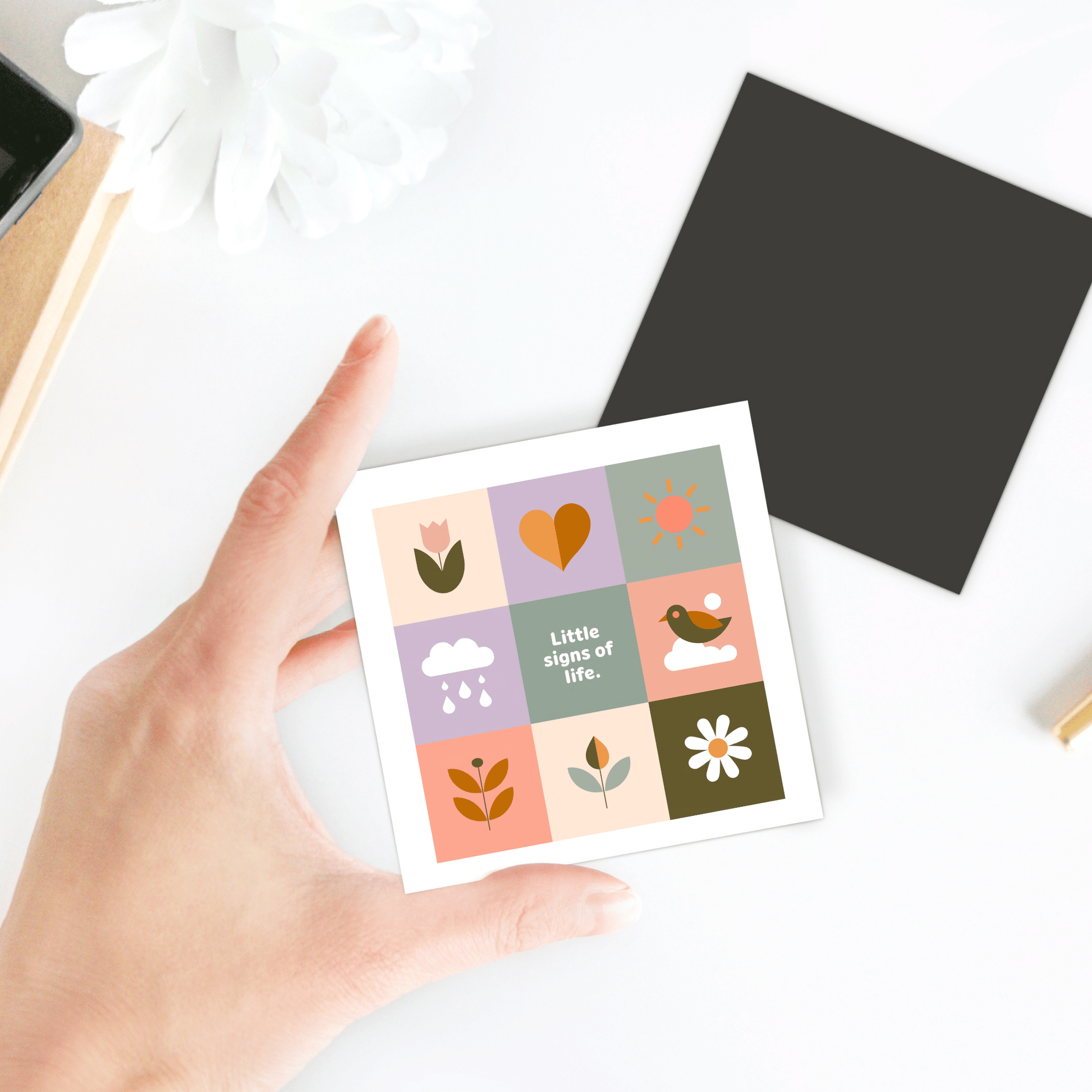 A hand holds the Mirabilia Boutique Little Signs of Life Pastel Grid Magnet, featuring colorful illustrations like a flower, heart, and sun with "Little signs of life" at center. A perfect inspirational gift or minimalist decor accent. Nearby are black and white squares.