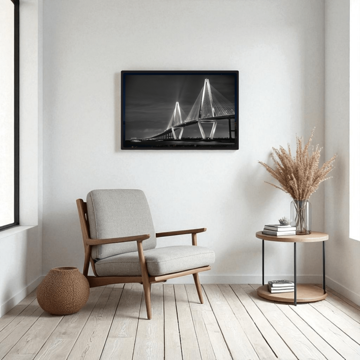 A modern, minimalist living room features a light armchair, round side table with dried plants, and the Ravenel Bridge Black & White Fine Art Print by Mirabilia Boutique on the wall. Light wood flooring and natural light complete the scene.