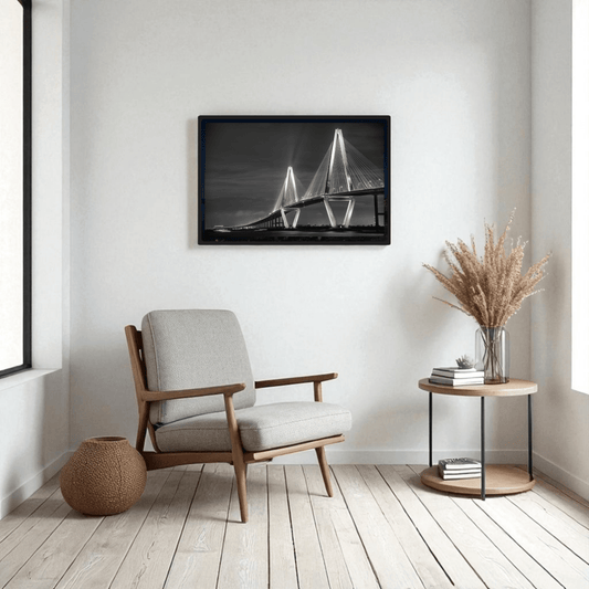 A modern, minimalist living room features a light armchair, round side table with dried plants, and the Ravenel Bridge Black & White Fine Art Print by Mirabilia Boutique on the wall. Light wood flooring and natural light complete the scene.