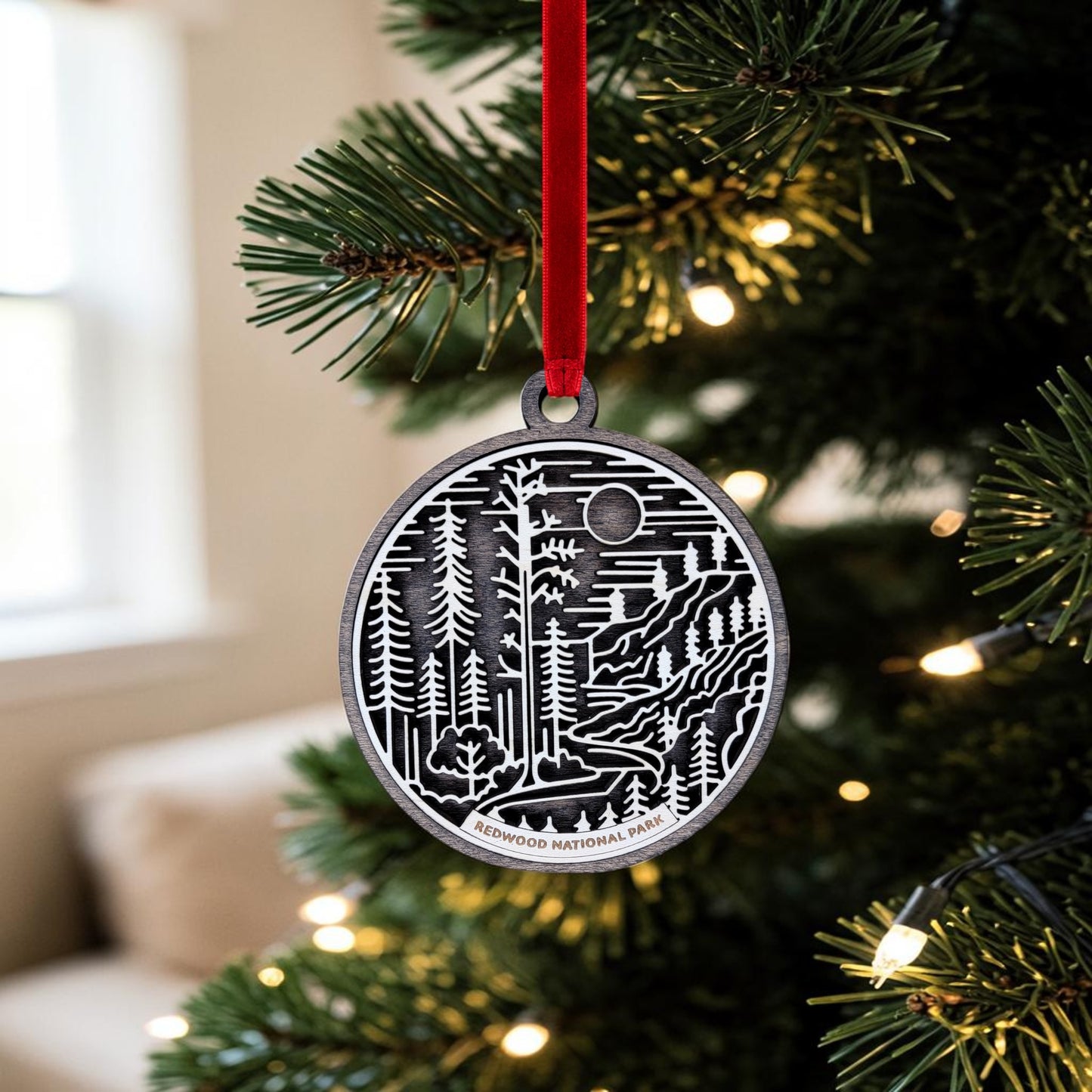 A Redwood National Park Line Art Christmas Ornament by Mirabilia Boutique, featuring intricate handcrafted layered wood with a forest design and “Redwood National Park” text, hangs from a red ribbon on a Christmas tree decorated with lights.