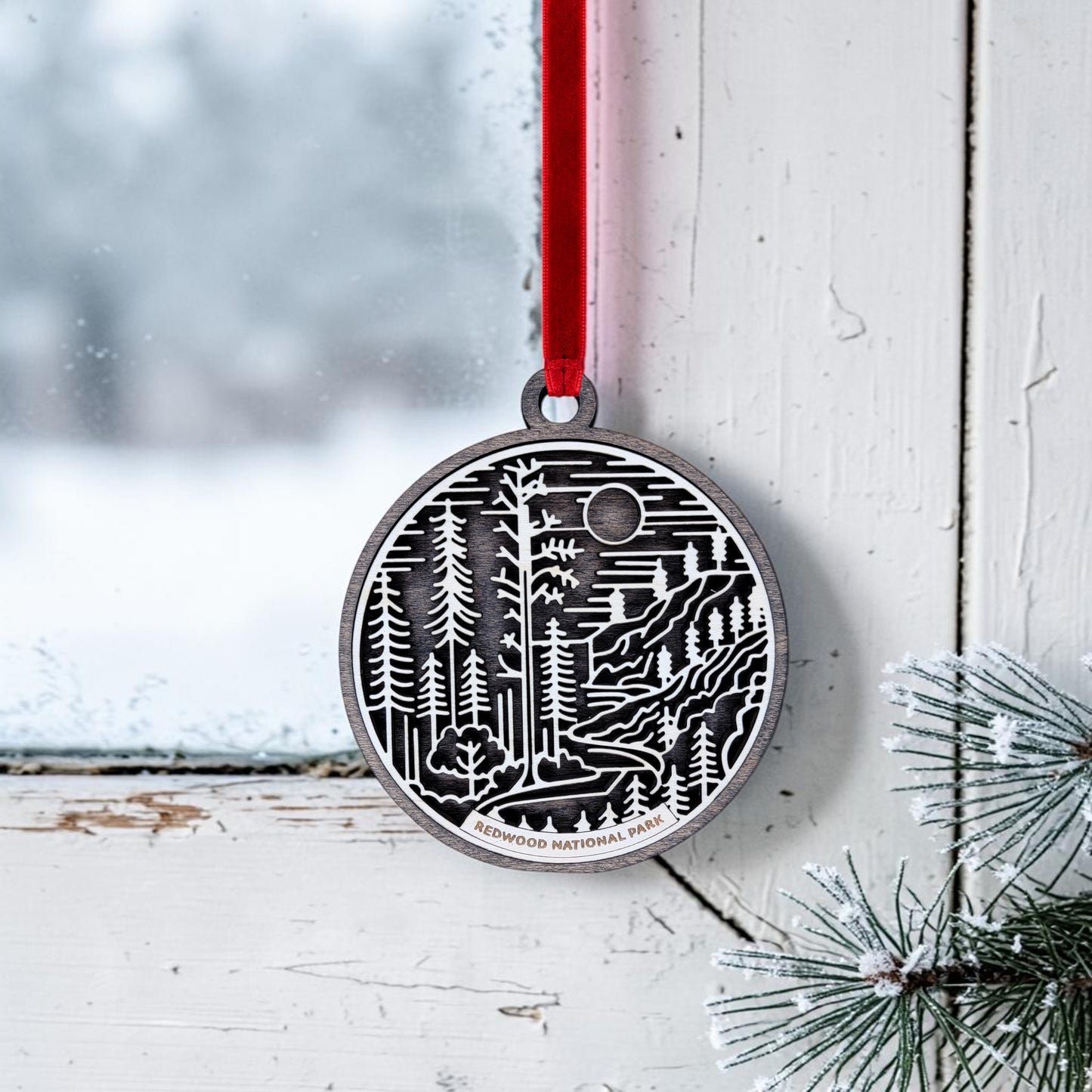 The Mirabilia Boutique Redwood National Park Line Art Christmas Ornament, a round silver piece featuring a layered forest design, hangs by a red ribbon on a white, frosted window. Snow-dusted pine branches frame the scene in the corner.