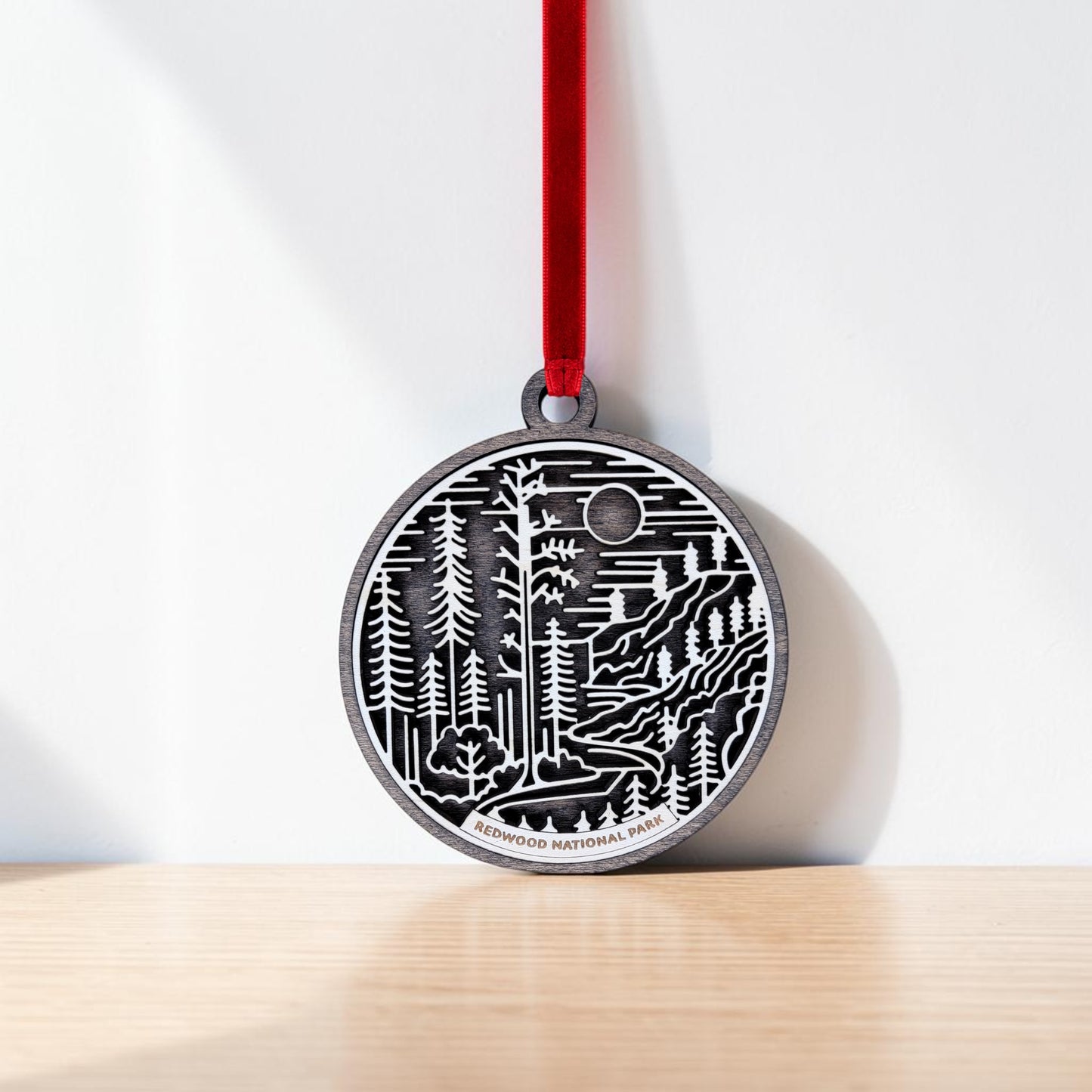 A round metal Redwood National Park Line Art Christmas Ornament by Mirabilia Boutique features engraved trees, a river, and sun in delicate layered line art. Hanging from a red ribbon, the ornament bears the text “Redwood National Park” at the bottom. It casts a shadow on a white wall above a wooden surface.