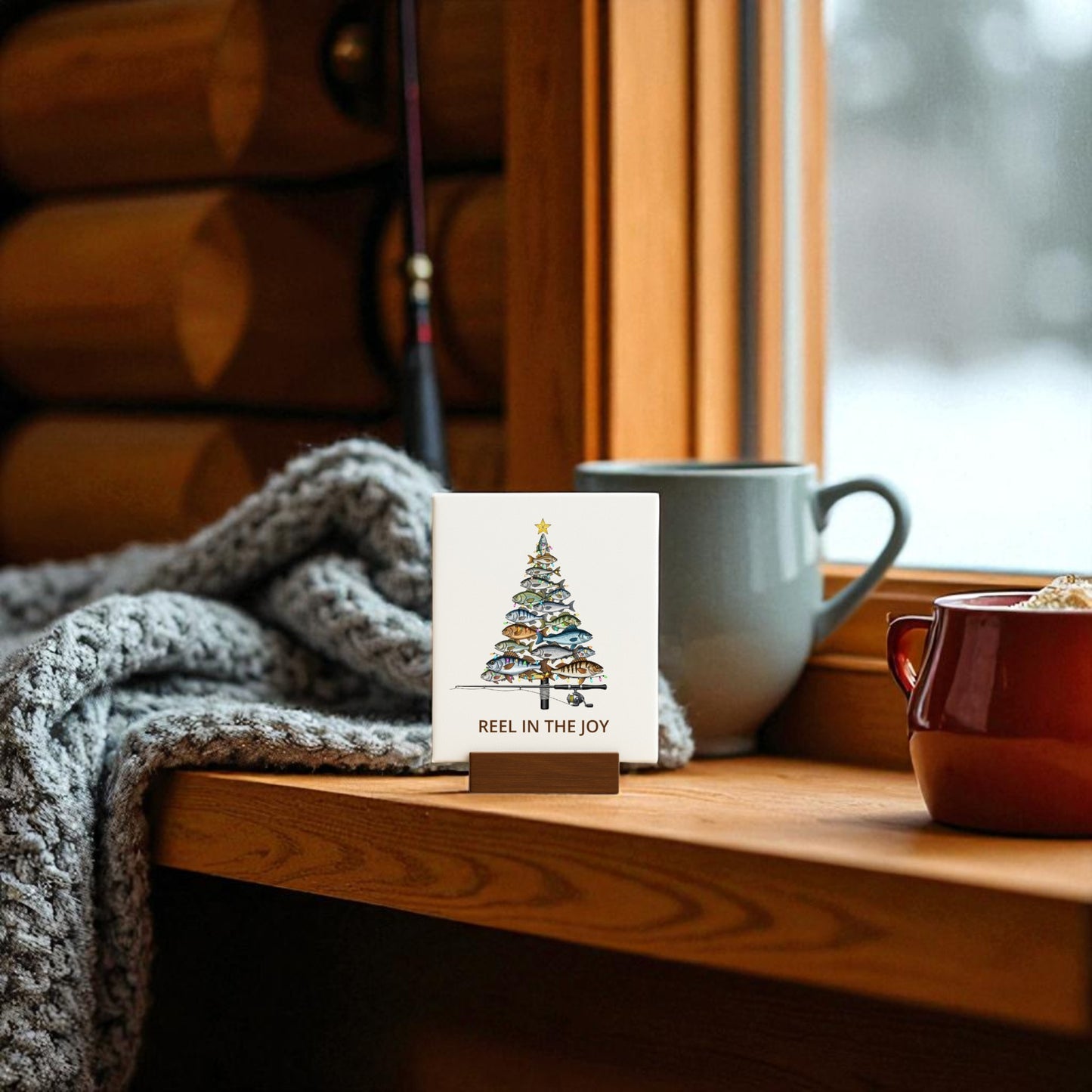 A cozy windowsill features Mirabilia Boutique’s Fishing Christmas Sign—"Reel in the Joy" canvas decor with a tree made of fish—plus a gray knitted blanket, blue mug, red cup, and softly falling snow outside.