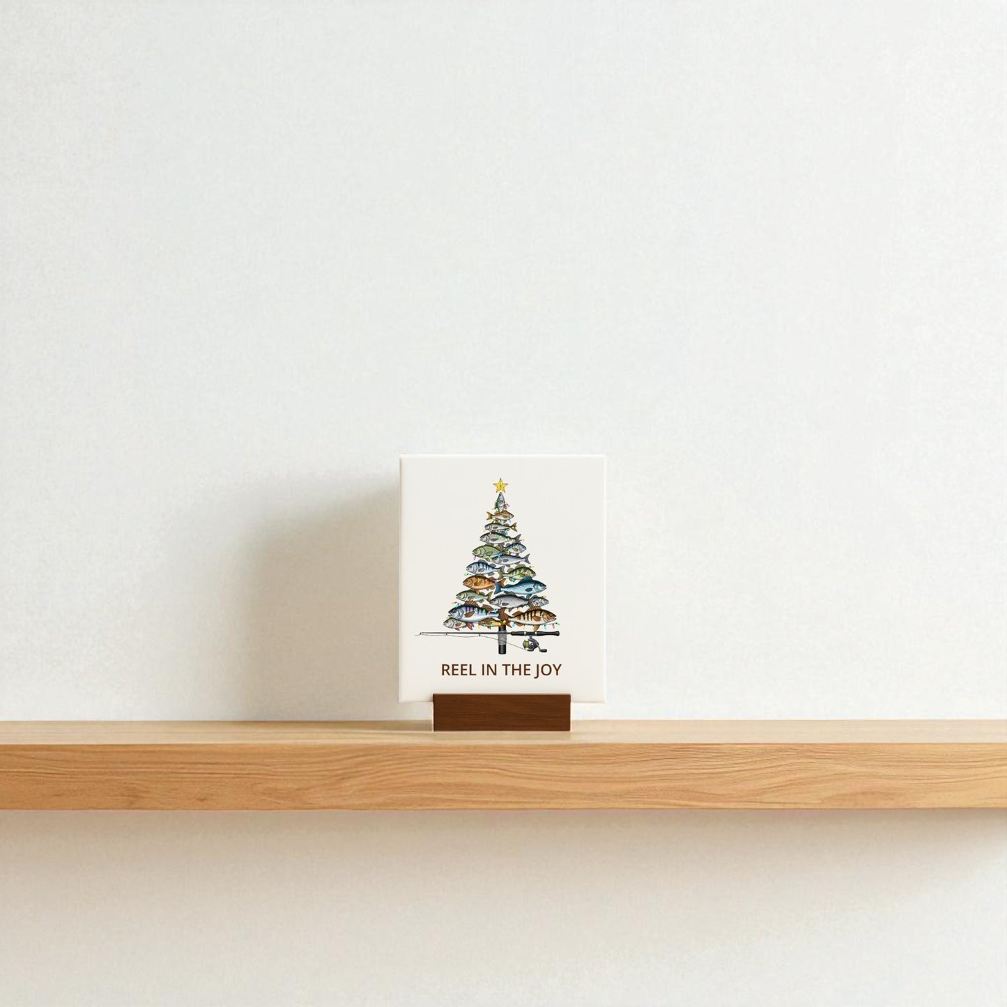 The Fishing Christmas Sign | Reel in the Joy Canvas Decor by Mirabilia Boutique features a fish-shaped Christmas tree and festive text, displayed on a wooden stand—perfect fishing-themed Christmas decor for any simple space.