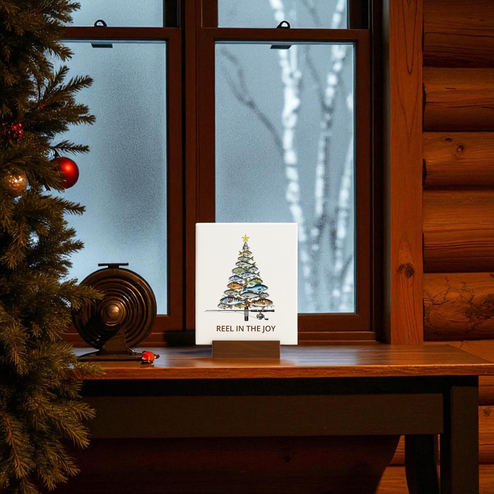 The Mirabilia Boutique "Fishing Christmas Sign | Reel in the Joy Canvas Decor" features a tree made of fish and festive text, displayed on a windowsill with a vintage camera, as snow falls outside.