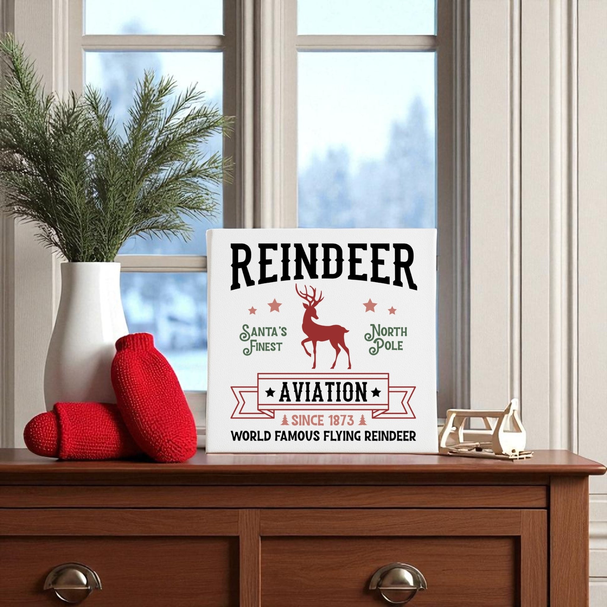 A vintage Christmas display features the Reindeer Aviation Christmas Sign by Mirabilia Boutique on a wooden table, paired with red knit mittens, a pine branch in a white vase, and a candle holder before large windows.