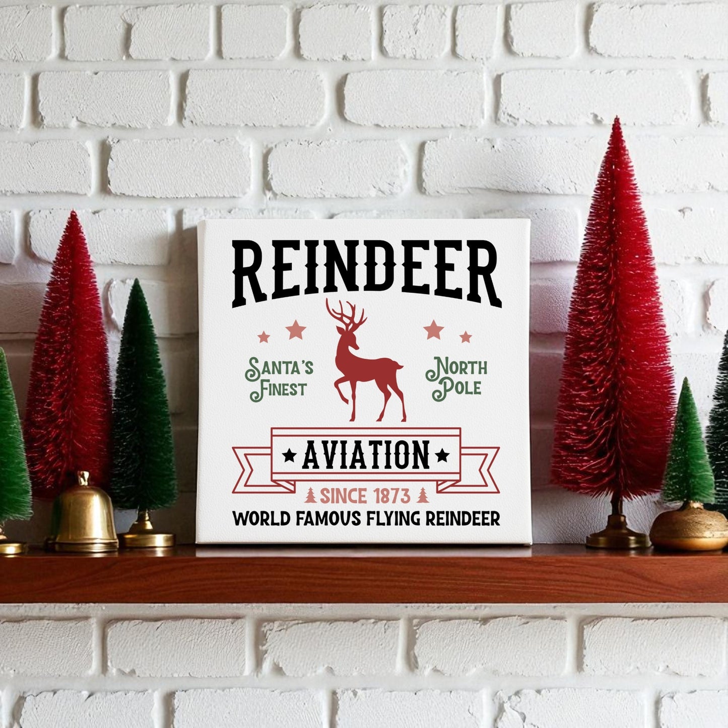 The Mirabilia Boutique "Reindeer Aviation Christmas Sign | North Pole Canvas Decor" sits on a white mantel, featuring a red reindeer and festive text. Red and green bottle-brush trees complete the vintage holiday scene.