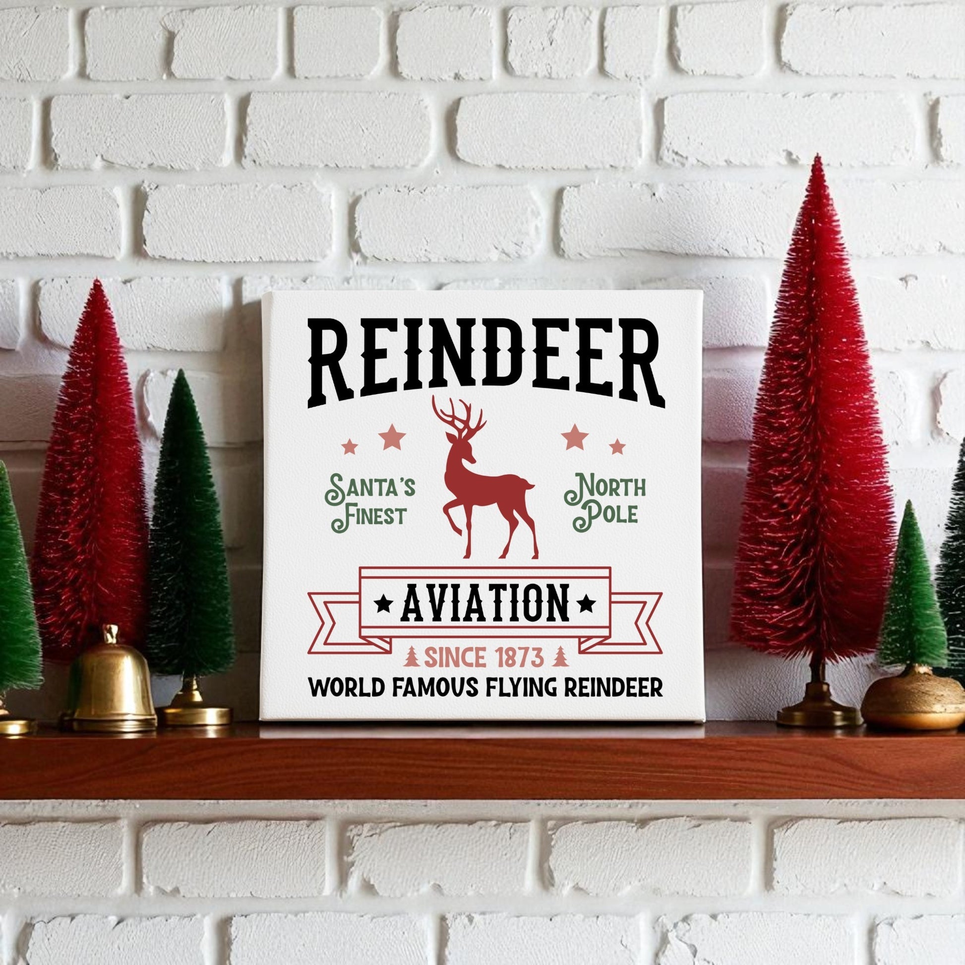 The Mirabilia Boutique "Reindeer Aviation Christmas Sign | North Pole Canvas Decor" sits on a white mantel, featuring a red reindeer and festive text. Red and green bottle-brush trees complete the vintage holiday scene.