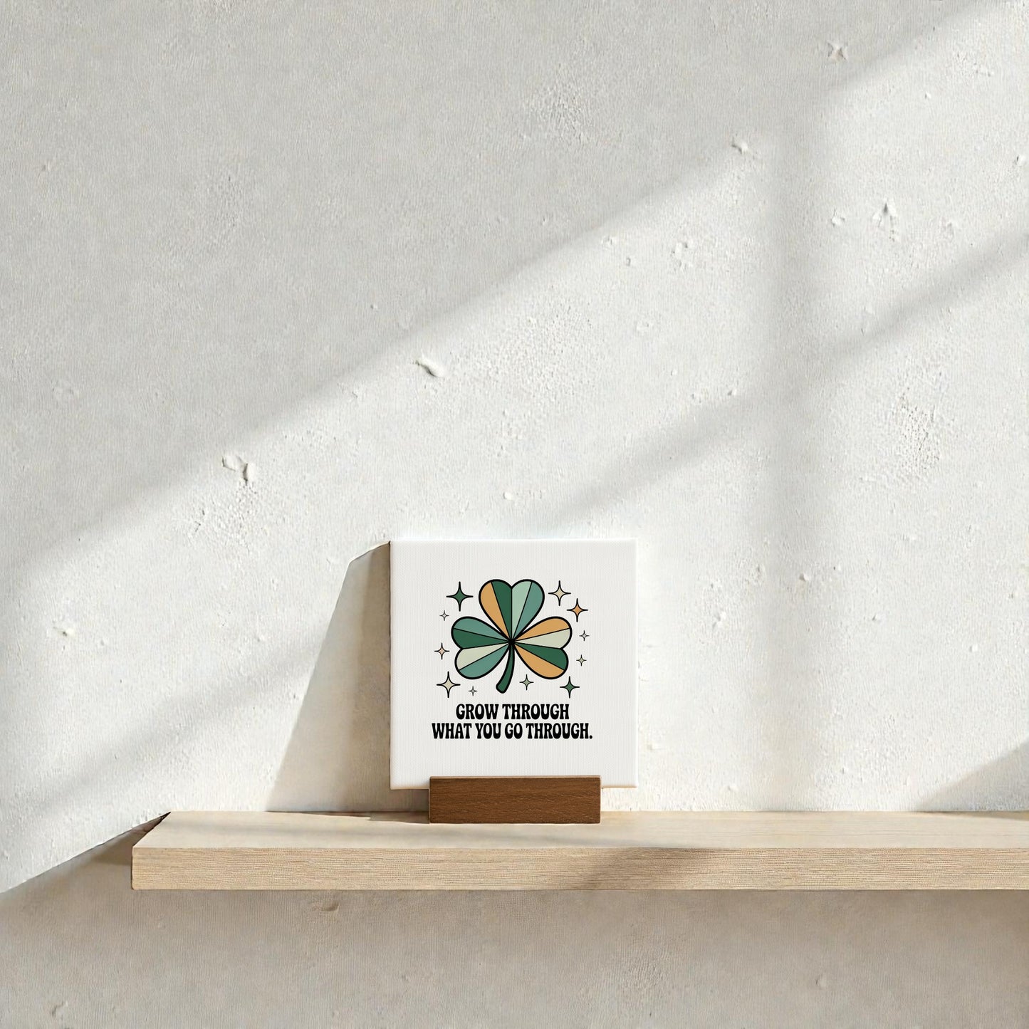 The Mirabilia Boutique "Grow Through What You Go Through" Mini Sign, featuring a green and brown four-leaf clover, sits on a sunlit wood shelf against a textured white wall.
