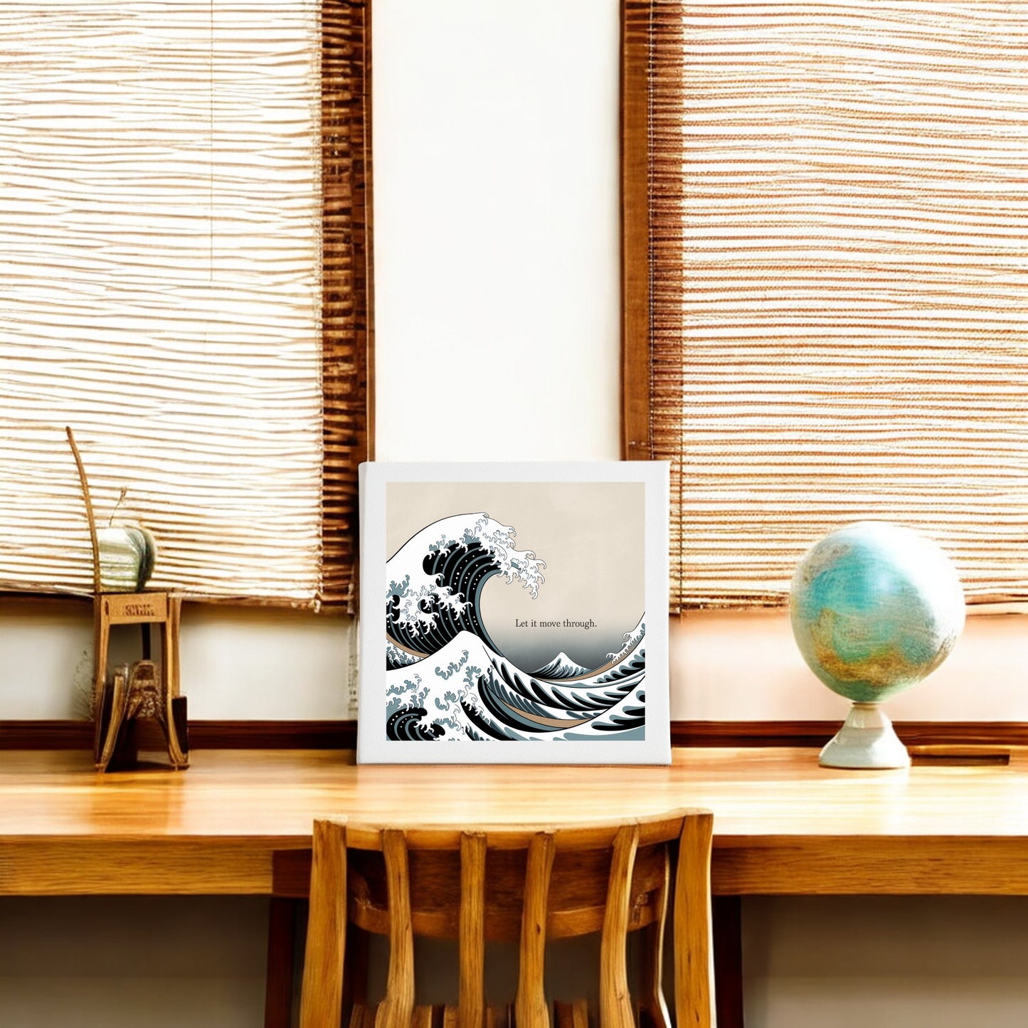 A wooden desk with a chair, a globe, and books sits by two large windows with woven blinds. Above is the Let It Move Through Canvas – Bold Ocean Calm Art by Mirabilia Boutique, featuring the phrase "Let it move through" and depicting emotional resilience.