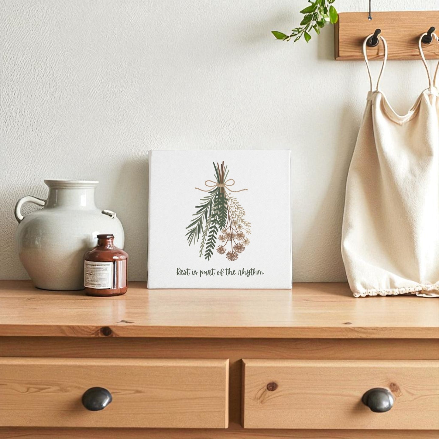 A wooden dresser features Mirabilia Boutique’s "Rest Is Part of the Rhythm" botanical canvas wall art, with a ceramic jug, small brown bottle, and a beige tote bag above, creating seasonal harmony in the space.