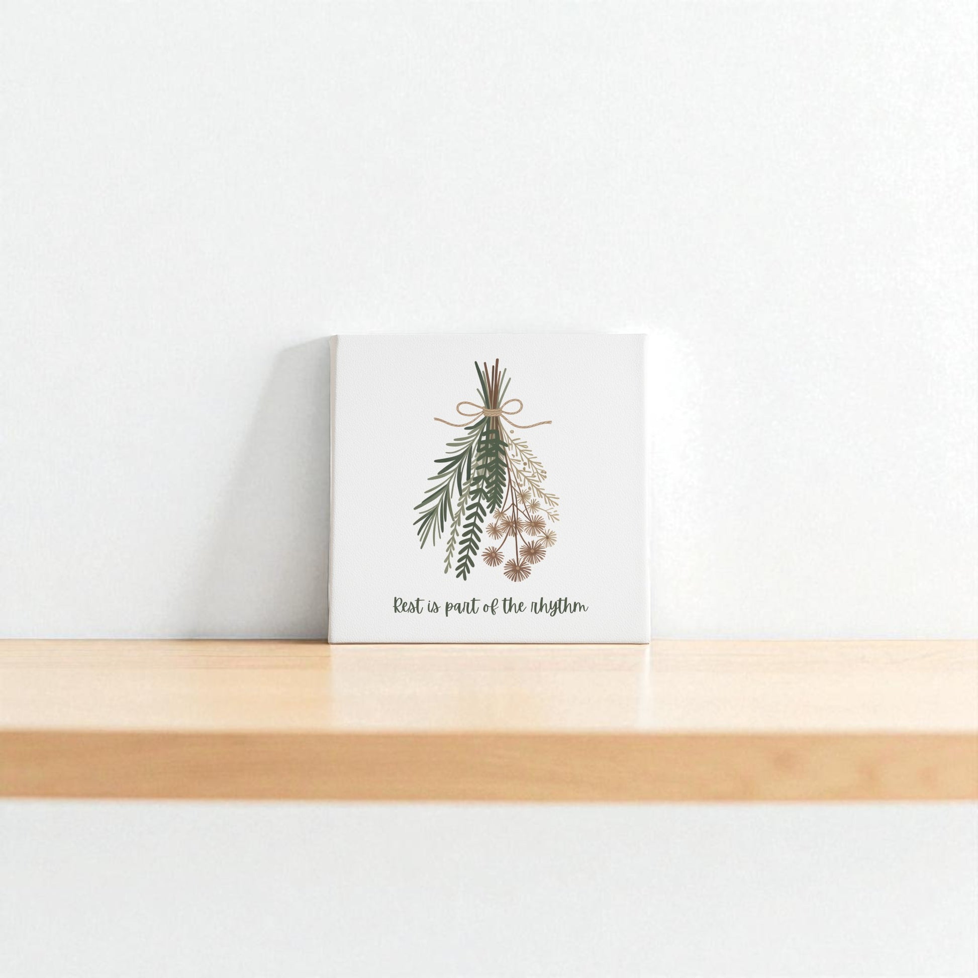 The Mirabilia Boutique “Rest Is Part of the Rhythm” Botanical Canvas Wall Art, a small minimalist square, sits on a light wooden shelf against a white wall, creating an atmosphere of seasonal harmony.