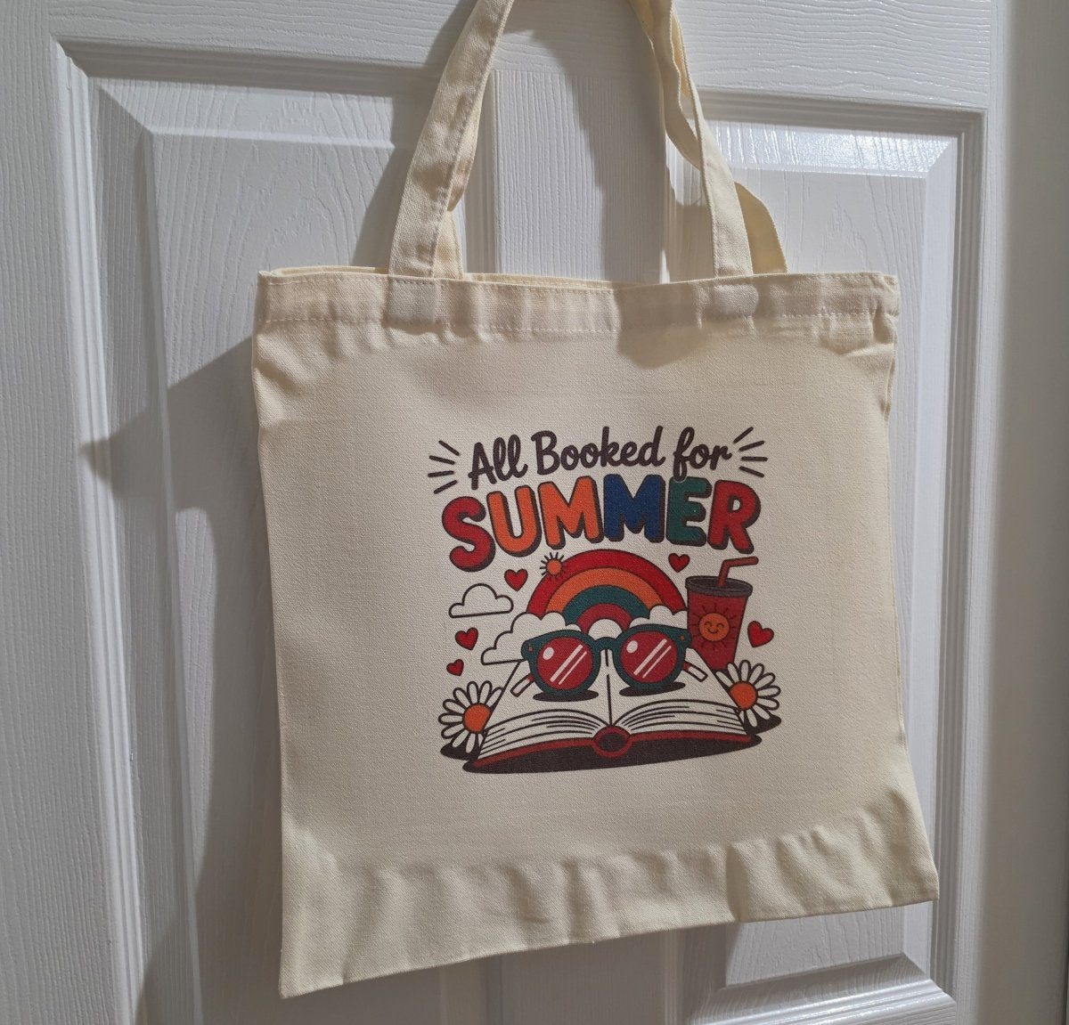 The Retro All Booked for Summer Tote Bag by Mirabilia Boutique is a cream cotton tote featuring a colorful design with a rainbow, sunglasses, an open book, drink, and playful “All Booked for SUMMER” text.