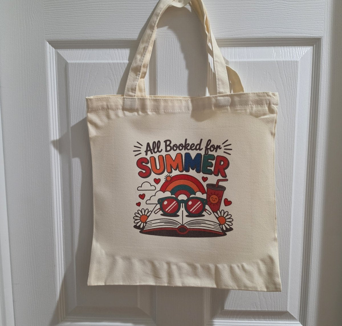 The Retro All Booked for Summer Tote Bag by Mirabilia Boutique hangs on a white door, featuring an open book, sunglasses, rainbow, sun, hearts, and “All Booked for Summer”—the perfect summer book tote.