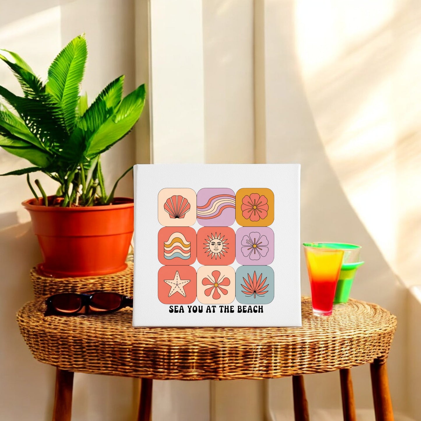 A rattan table holds sunglasses, a tropical drink, and a plant beside Mirabilia Boutique's Sea You at the Beach Canvas – Retro Coastal Wall Art. Sunlight enhances the relaxed retro summer vibe.